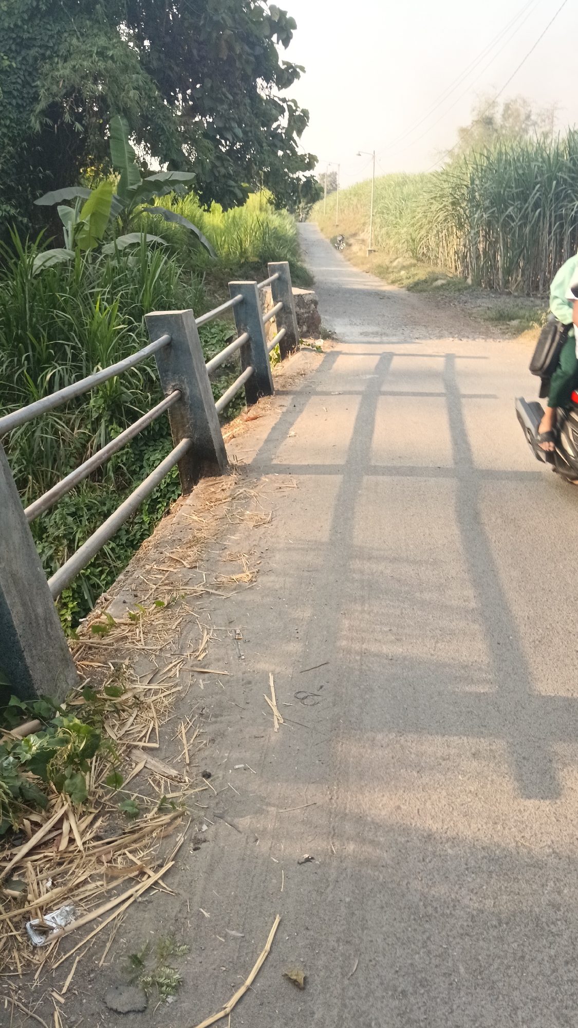 The image shows a dirt road bordered by lush vegetation and tall sugarcane fields. The road leads uphill, with a slight curve to the left. A motorcycle is visible in the distance, and the shadow of a guardrail is cast across the road. The scene appears to be located in the city of Slumbung, Indonesia, based on its rural setting and the presence of sugarcane. The time of day is likely late afternoon, given the long shadows. The overall impression is one of a peaceful, rural landscape.