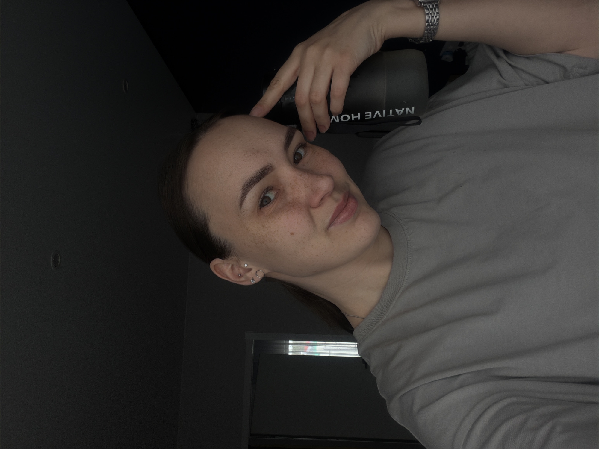 A woman with freckles is holding a dark-colored bottle with the words "NATIVE HOME" printed on it. She is wearing a gray shirt and a silver wristwatch. She is indoors, and part of a window and a dark gray wall are visible in the background. Her location in the city of Chaikovsky, Russia, cannot be determined from the image.