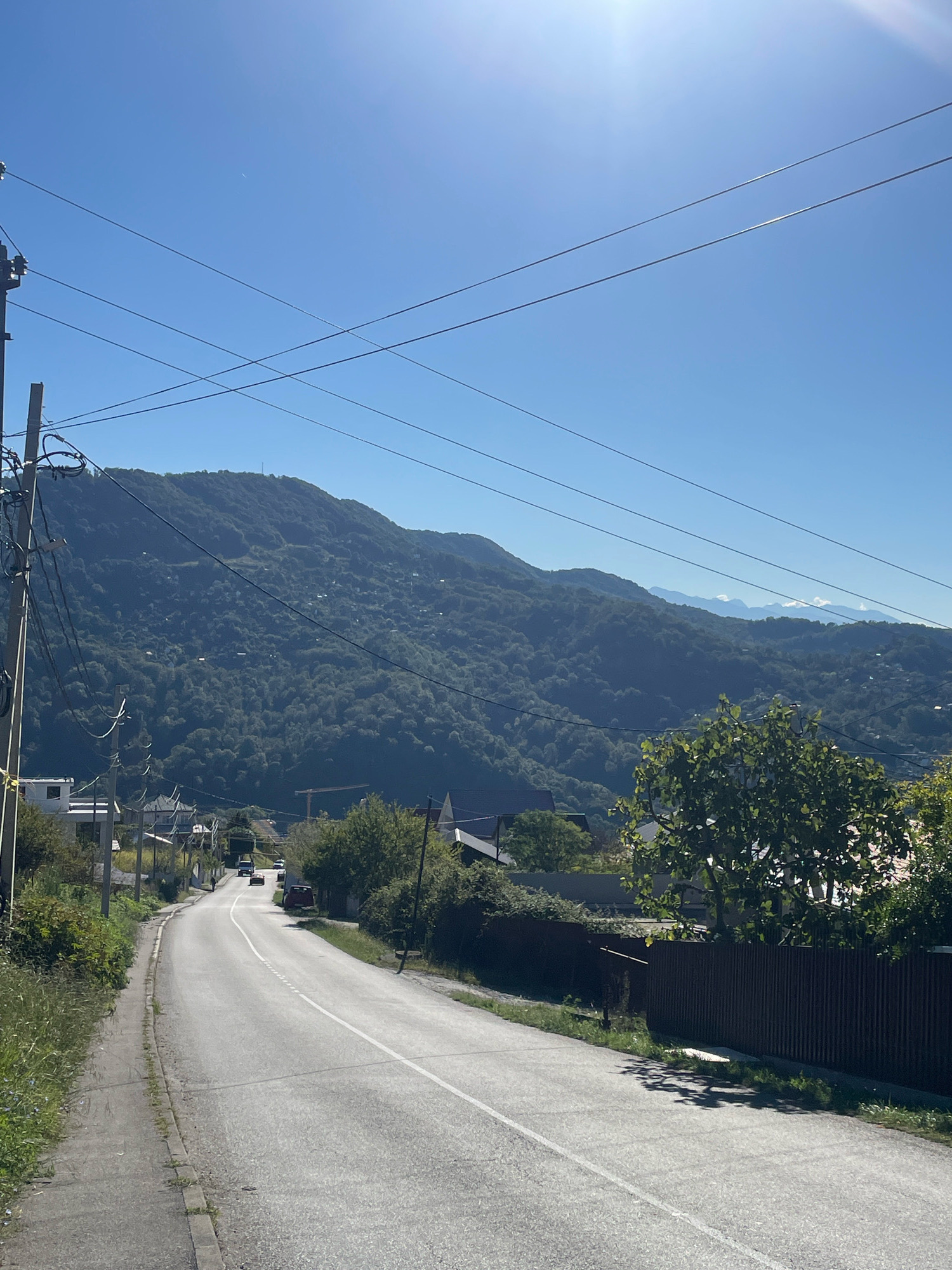 The image depicts a paved road winding uphill through a rural or suburban area. The road is bordered by a grassy verge on the left and a dark, corrugated fence on the right. Several vehicles, including cars and a van, are visible on the road. Buildings and trees are scattered on the slopes leading up to a large, heavily forested mountain range that dominates the background. The sky is clear and bright blue, with a subtle lens flare from the sun in the upper right quadrant. Overhead power lines crisscross the sky. The scene appears to be set in the city of Baranovka, Russia, given the context provided. The overall impression is one of a sunny day in a mountainous landscape with scattered human habitation.
