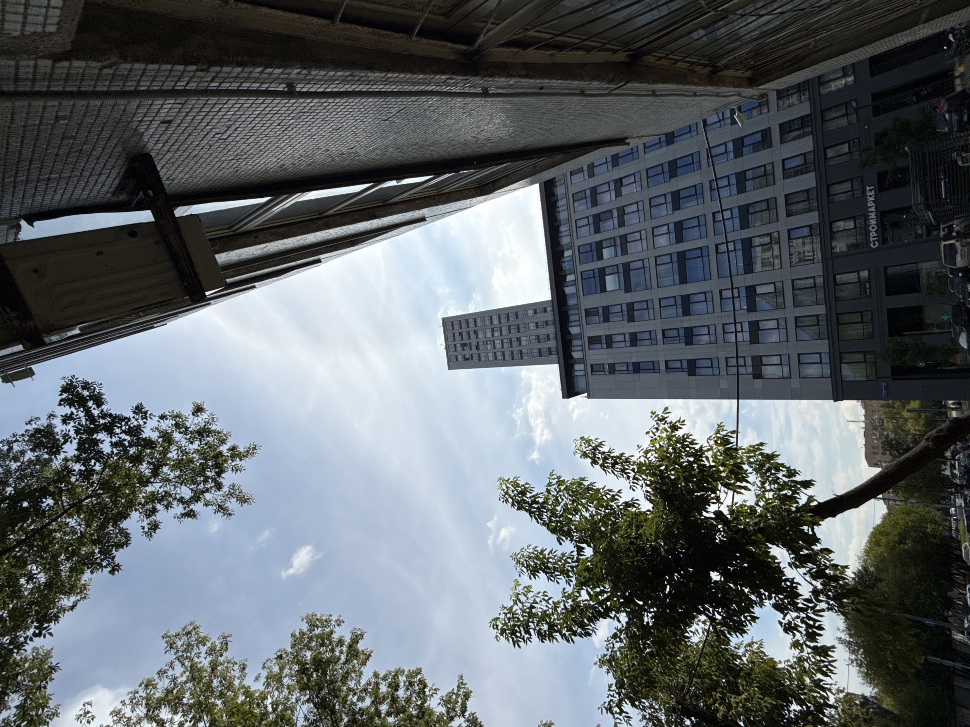 The image, taken from a low angle looking upwards, captures a street scene in Moscow, Russia. The foreground features the textured facade of a building on the right, with visible brickwork and windows. In the center, a modern, tall grey building with numerous windows dominates the view, and a much taller, slender skyscraper stands further in the background. To the left, lush green trees with dense foliage are visible against a bright blue sky with scattered white clouds. Below the trees, hints of a street with parked cars and trees can be seen. A sign on the lower part of the central building reads "СТРОЙМАРКЕТ" (StroyMarket), indicating a construction materials market. The overall atmosphere is that of an urban environment on a clear day.