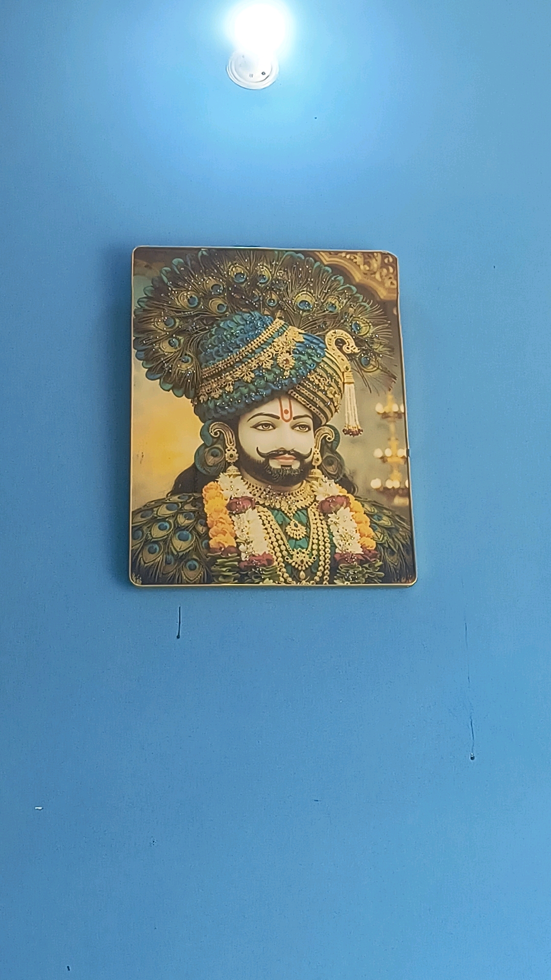 The image shows a portrait of a man adorned in a richly decorated turban and traditional Indian attire, featuring intricate peacock feather motifs. The man has a prominent mustache, a beard, and is wearing several necklaces. He has a red mark on his forehead. The portrait is framed and hangs on a vibrant blue wall. Above the portrait, a white ceiling light is visible. The photograph appears to have been taken indoors, likely in a home or a place of worship, in Loni, India.