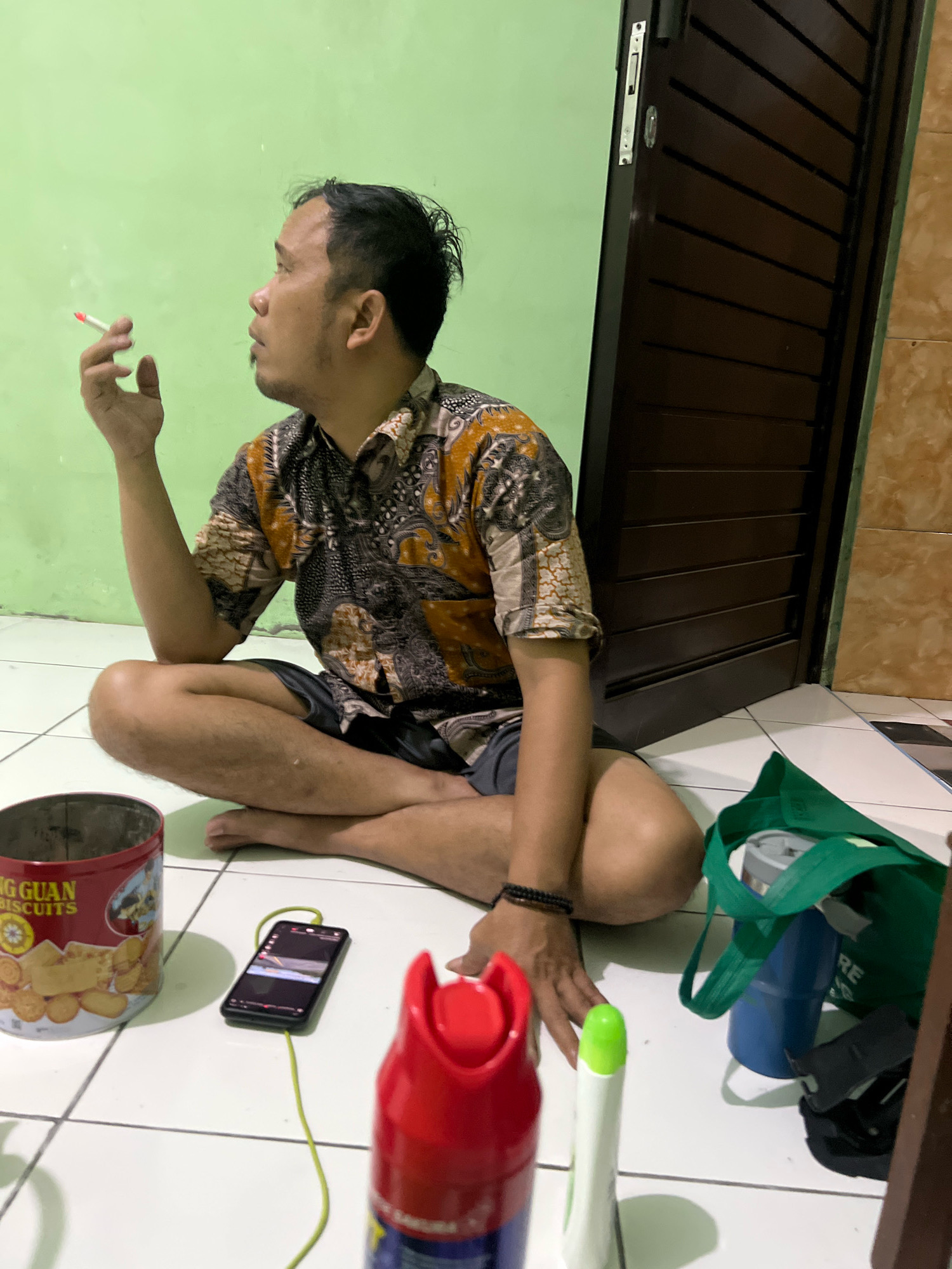 A man is sitting cross-legged on a tiled floor in what appears to be an indoor space with a light green wall and a dark brown door. He is wearing a patterned short-sleeved shirt and dark shorts. The man is holding a lit cigarette in his right hand and looking to his left. In front of him on the floor are several objects: a red tin can labeled "NG GUAN BISCUITS", a smartphone connected to a charging cable, a red spray can, a white spray can with a green top, and a green tote bag containing a blue bottle and other items. The lighting suggests it is daytime. The scene is likely a residential setting in Tlogoadi, Indonesia.