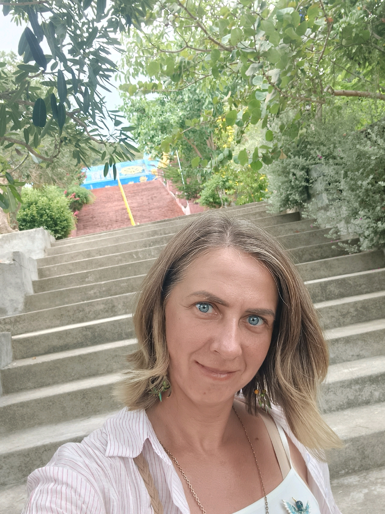 A woman with blonde hair and blue eyes is taking a selfie on a set of outdoor concrete stairs. She is wearing a white shirt with pink stripes and a white tank top underneath. She has on a gold chain necklace with a bird-shaped pendant. Behind her, the stairs lead up to a red-tiled structure with blue and yellow decorations at the top. Lush green trees and foliage surround the staircase. The location is the Long Son Pagoda in Nha Trang, Vietnam.