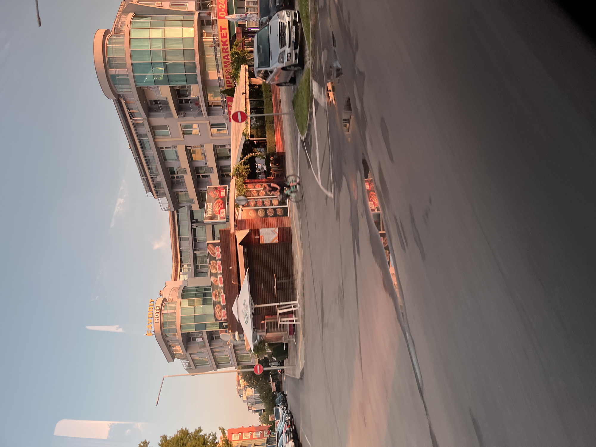 The image depicts a street scene in Nesebar, Bulgaria, at sunset. A modern, multi-story building with curved sections and glass balconies dominates the background. A sign on the building reads "FAVORIT HOTEL". In front of the hotel, there is a strip of businesses, including a food stall with a "Pizza" sign and another with a "SUPERMARKET 0-24" sign. A person is riding a bicycle on the street in front of the food stall. Several cars are parked along the street and on the side of the road. Puddles of water on the asphalt street reflect the sunset sky. The overall atmosphere suggests a warm, late afternoon or early evening in a coastal town.