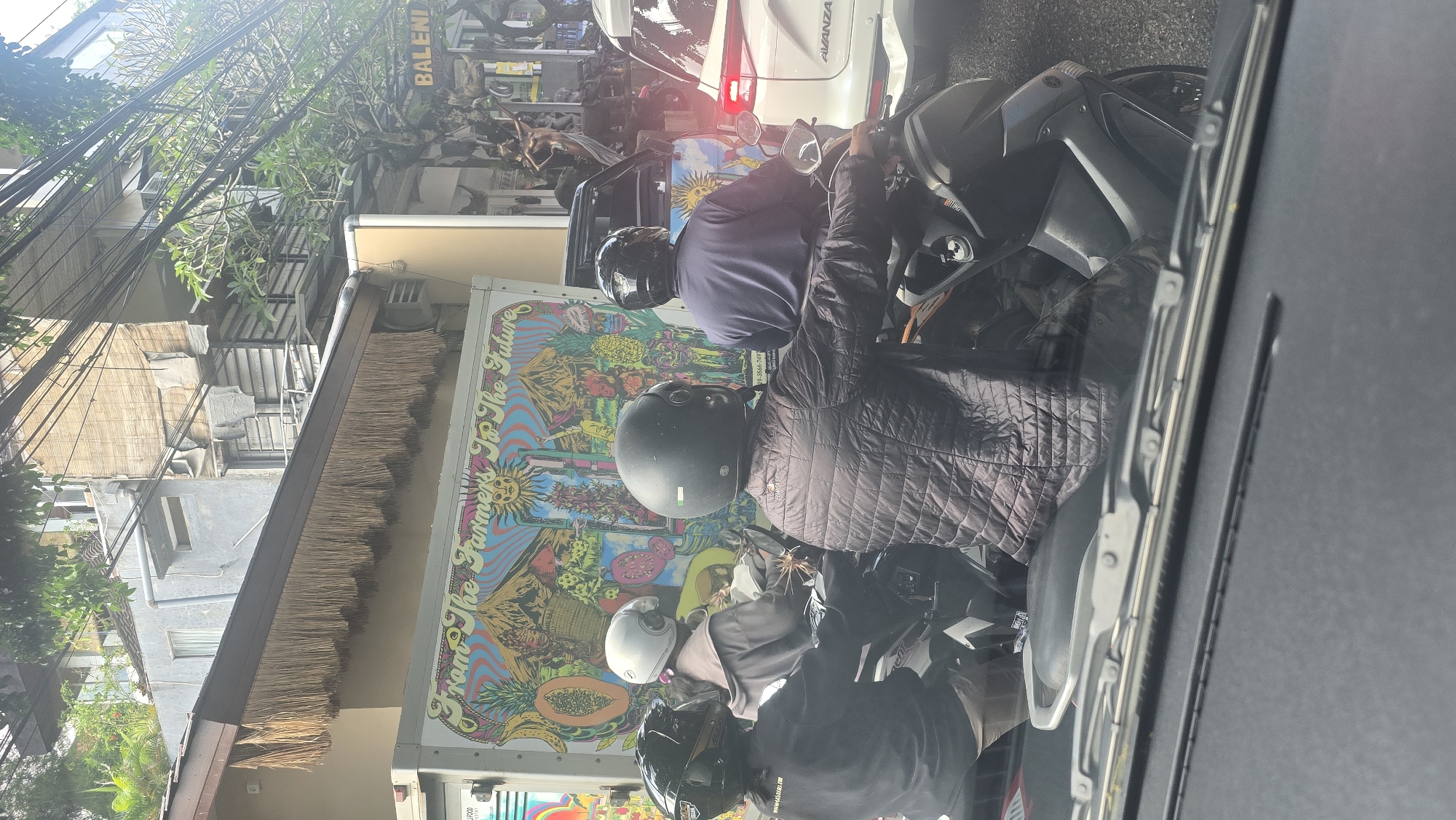 The image captures a scene in Ubud District, Indonesia, as viewed through the windshield of a vehicle. Several people on motorbikes are waiting in traffic. The person closest to the camera is wearing a black helmet and a dark quilted jacket. The driver of a white minivan is visible. The side of the van has an artistic mural promoting "Grow The Farmers To The Future". Overhead electrical wires are visible near buildings. The lighting suggests it is daytime.