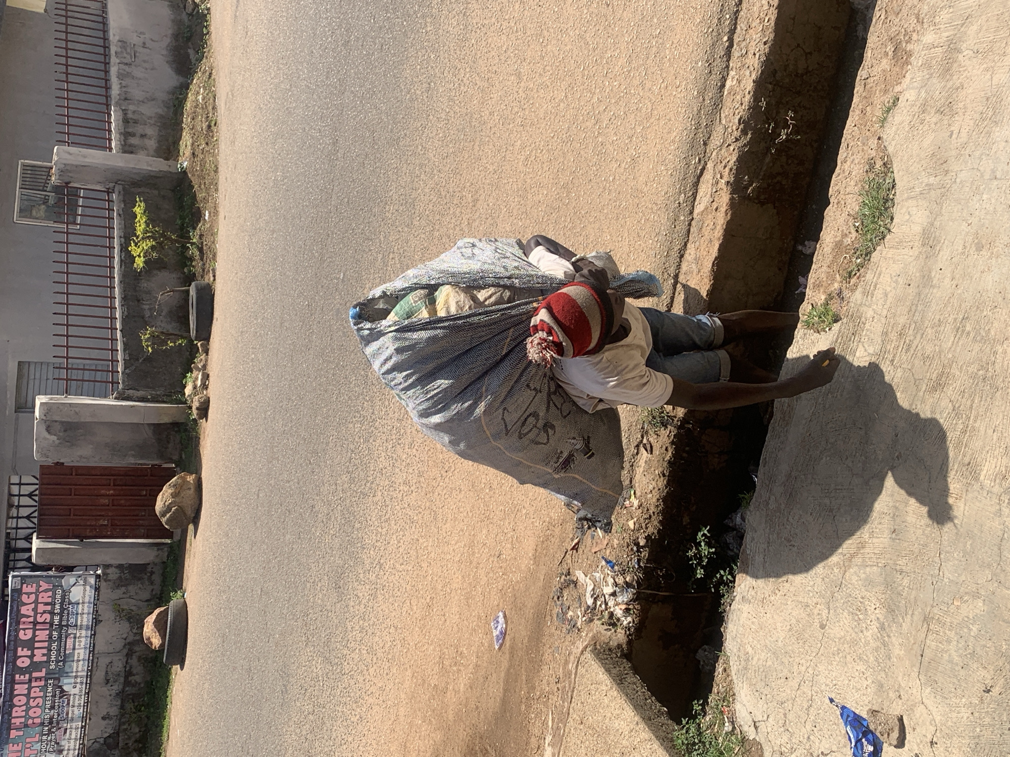 An image captured in Jos, Nigeria, shows an individual resting beside a large, bulky sack on the side of a paved road. The subject, appearing male, is wearing a white t-shirt, blue jeans, and a red and white striped beanie, and is leaning against the large, patterned (grey and white) sack. The sack appears full, with light-colored contents visible at the top, possibly collected materials.
The scene depicts a street environment under bright, sunny conditions, indicated by strong shadows cast across the ground. The weather appears clear. To the left, there's a grey building with a metal fence and gate, and a window. Sparse vegetation, including small plants and grass, grows in a drainage ditch running parallel to the road and a concrete sidewalk. Several rocks and an old tire are visible along the roadside and near the building, possibly used as makeshift barriers. Some litter is scattered in the ditch and on the road.
A partially visible sign on the building's wall includes text for "THE THRONE OF GRACE INTERNATIONAL GOSPEL MINISTRY," advertising "HOUR IN HIS PRESENCE" (Prayer & Intercession) at 5:00 PM and "SCHOOL OF THE SWORD" (A Community Bible Class) on Thursdays at 4:30 PM. Contact information like "@The ThroneofGracelet Gospel," "throneofgrace2025," and a phone number (+234-704-365-6857) are also present, along with an address mentioning "ITF Corpers Lodge, Dadin Kowa."