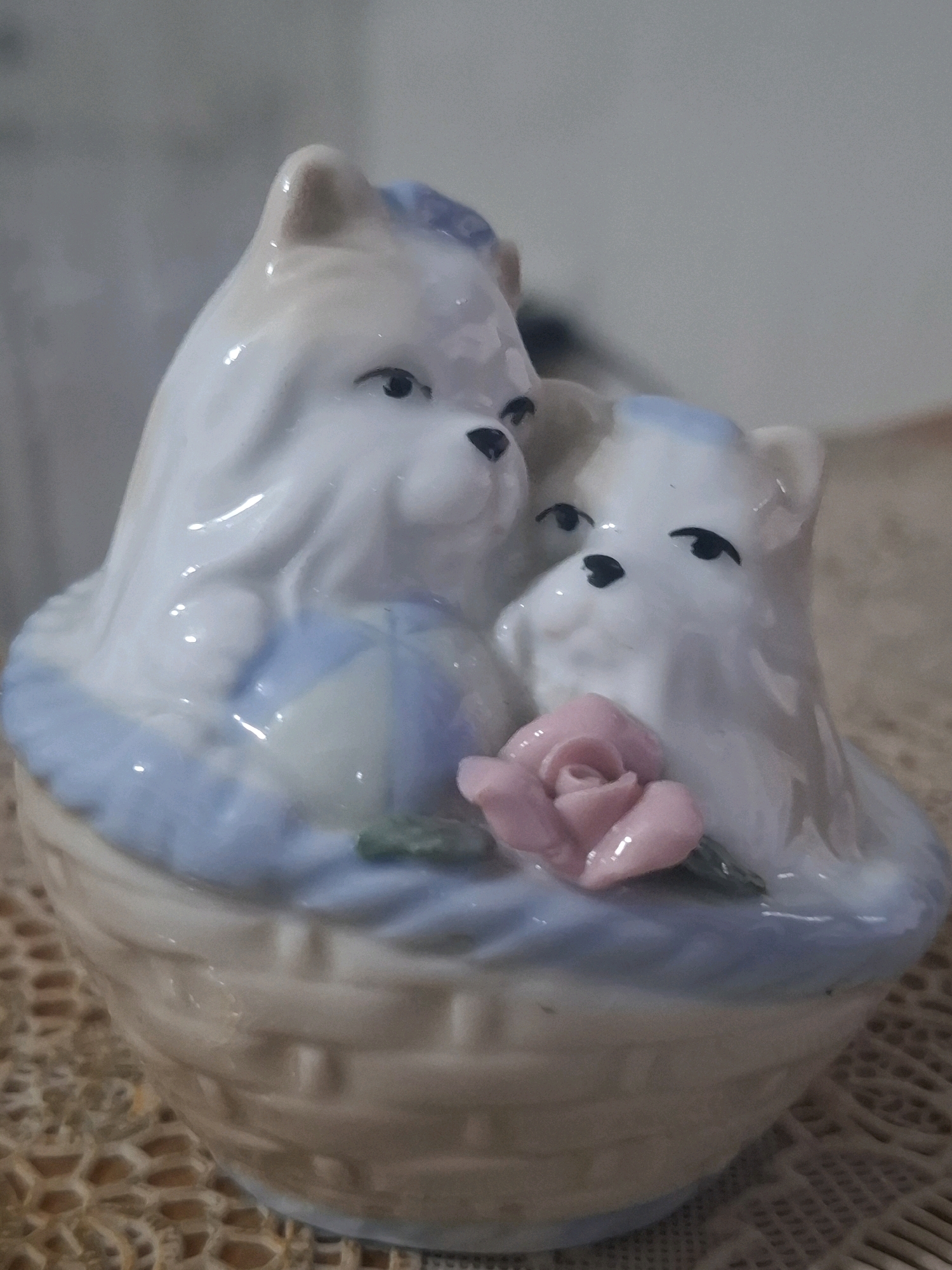 A ceramic figurine of two white dogs in a basket sits on a lace doily. The dogs are depicted with light fur, blue accents on their heads, and black eyes and noses. One dog is larger than the other, and they appear to be nestled together. A small pink rose with green leaves is placed in front of them. The basket has a woven texture and a light blue rim. The background is blurred and appears to be an indoor setting with neutral-toned walls. There is no identifiable information that places this image in Almaty, Kazakhstan.