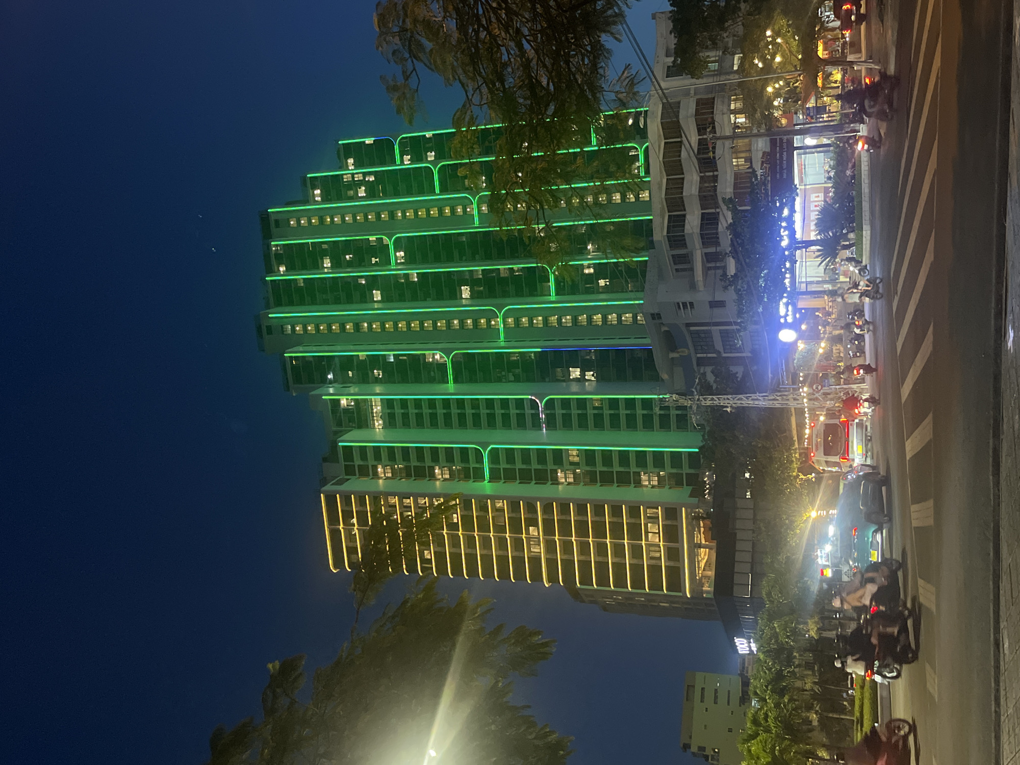 The image shows a night scene in Bac Nha Trang Ward, Vietnam. A tall building dominates the left side of the frame, illuminated with green lights on the upper floors and yellow lights on the lower floors. A street runs along the right side of the building, with various vehicles including cars and motorcycles. Pedestrians and trees line the street. Streetlights provide additional illumination. Other buildings are visible in the background. The sky is dark.