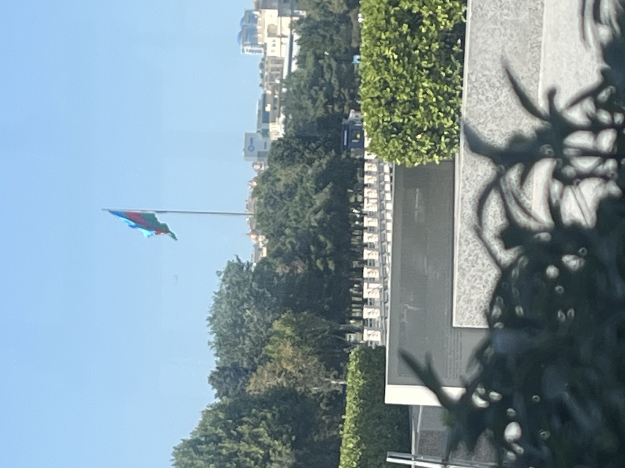The image depicts a view from an elevated position, looking out over a manicured garden area with lush green bushes and trees. In the background, several buildings are visible, including a modern building with blue accents and other structures that suggest an urban environment. A flag, which appears to be the flag of Azerbaijan (three horizontal bands of blue, red, and green, with a white crescent and star in the center), is prominently displayed on a tall flagpole. The sky is clear and blue. In the foreground, there is a blurred view of dark green foliage, possibly from plants or trees, obscuring parts of the scene. A white rectangular structure with what appears to be engraved text is also visible in the middle ground, partially obscured by the foreground foliage. The overall impression is that of a well-maintained public space or memorial in Baku, Azerbaijan, possibly with a view of the city skyline.