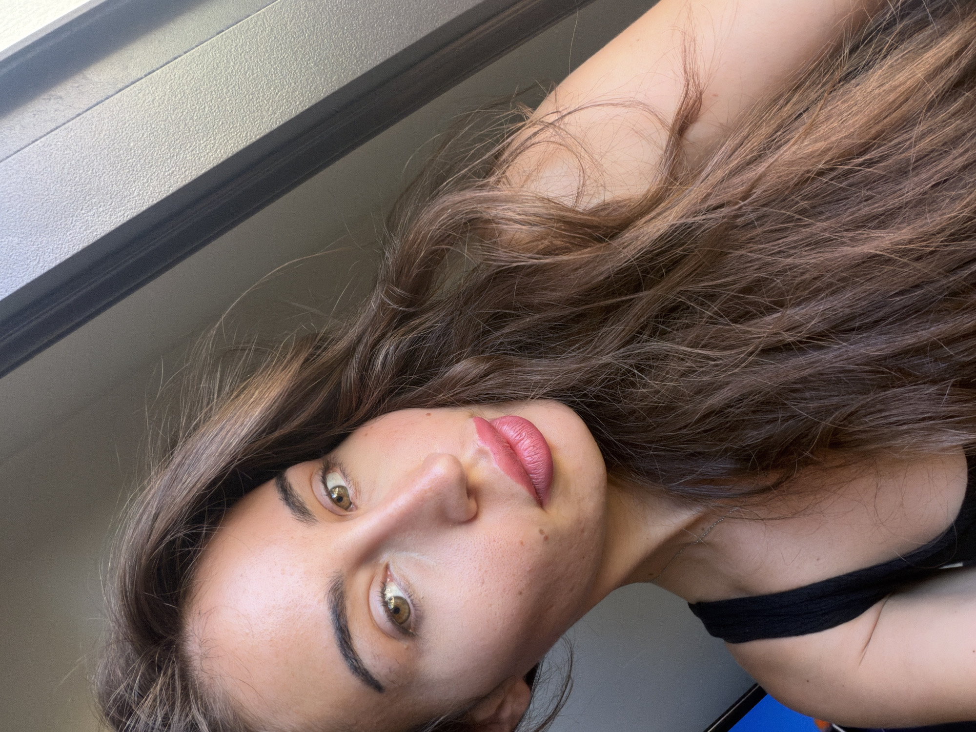 A medium shot photo of a woman with long, wavy, brown hair is captured from the shoulders up. She is looking towards the camera with a neutral expression. Her makeup includes pink lipstick and eye makeup. She is wearing a black tank top. The background is out of focus and appears to be an indoor setting with natural light coming from the side, possibly near a window or door frame. There is a vertical element with a textured surface on the right side of the frame, which is in shadow and light. The image has a warm tone, indicating natural lighting. The location of Tersefanou, Cyprus, is not evident from the visual cues in the image itself.