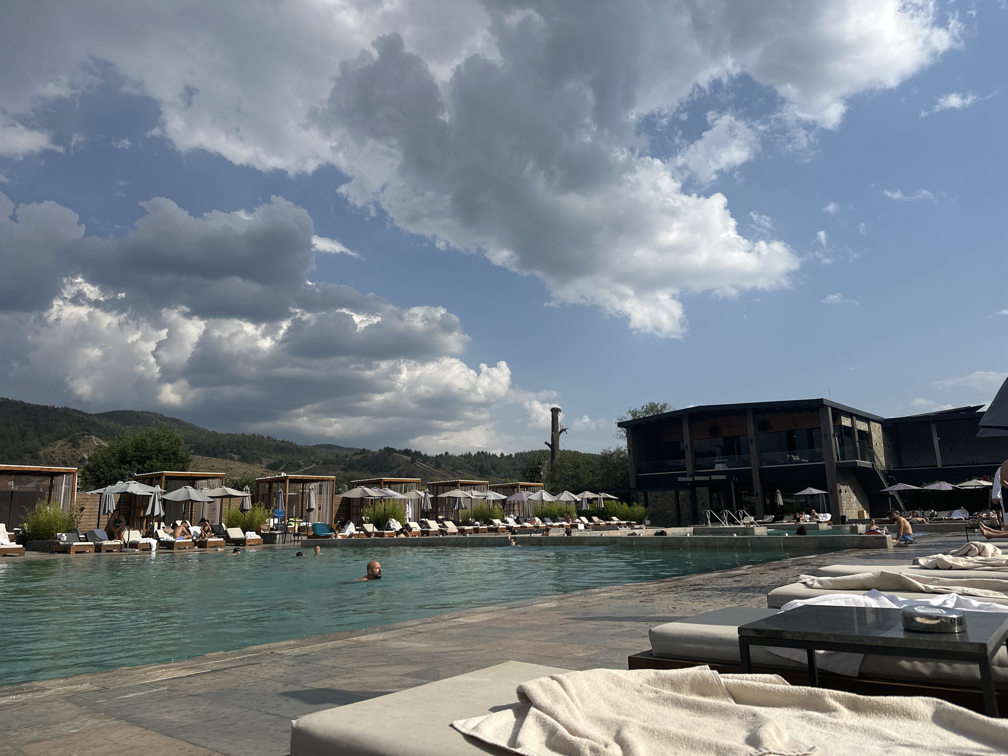 In Bania, Bulgaria, an outdoor swimming pool area is depicted under a partly cloudy sky. The pool itself is large and filled with clear turquoise water. Several people are visible in the water, with one man in the foreground swimming. Around the pool, there are numerous lounge chairs, some occupied by people relaxing or sunbathing, and others are empty. Many of these chairs are shaded by umbrellas or covered cabanas with tan fabric. In the background, a modern, dark-colored building with multiple levels and large windows is situated. Beyond the building and pool area, rolling hills covered with trees and vegetation rise, with a few scattered structures visible on their slopes. The overall scene suggests a resort or hotel complex designed for leisure and relaxation.