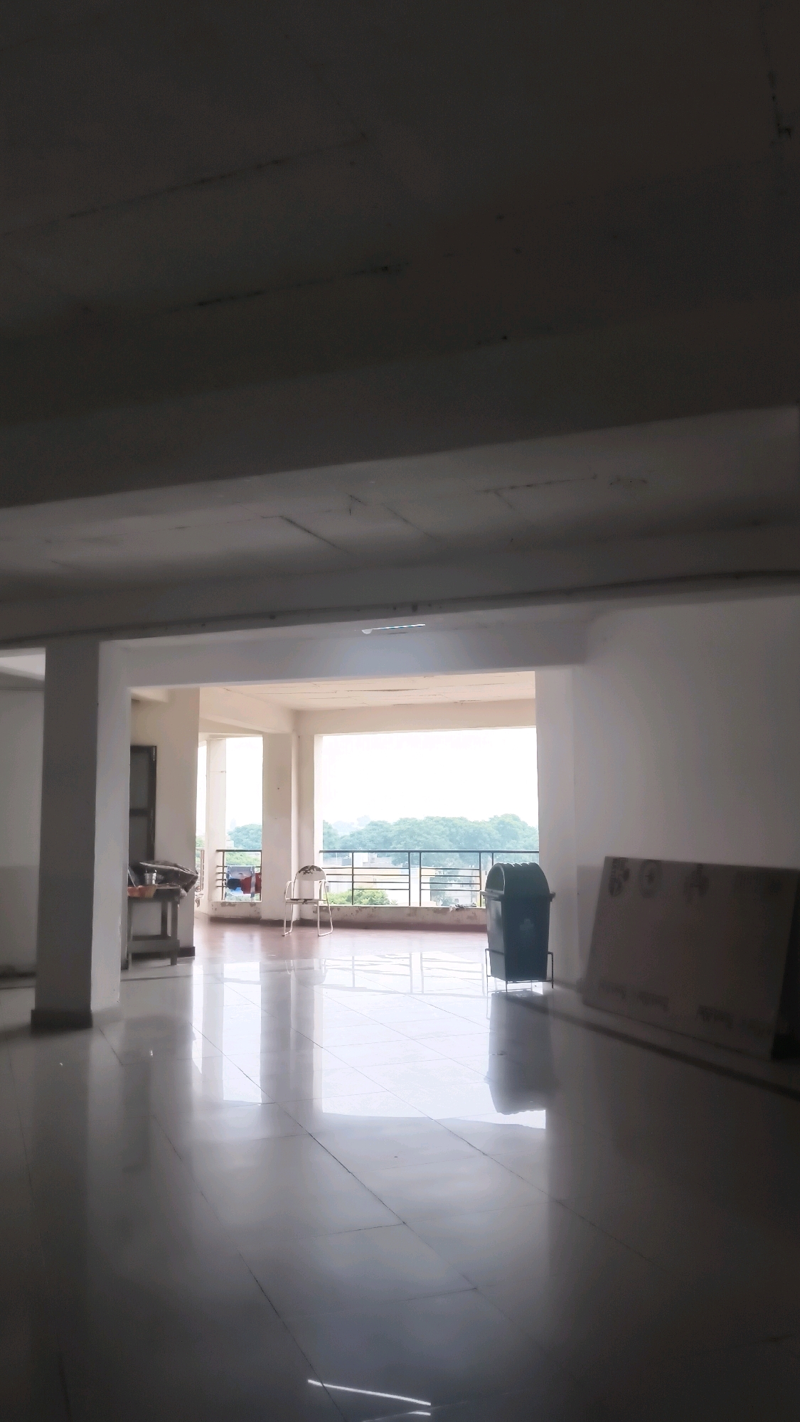 A wide shot captures the interior of a spacious, unfinished concrete structure, possibly a building under construction or renovation, with a view of lush green trees and a hazy sky through an open balcony. The floor is a polished, reflective surface, marked by a grid of square tiles that mirror the overhead concrete ceiling and the ambient light. Several white concrete pillars support the ceiling, dividing the space.
To the left, a small wooden table holds a few items, partially obscured by shadow. Further to the left, a tall, dark doorway is visible. In the center of the frame, a single, metallic folding chair stands on the balcony, facing outwards. Next to the chair, a dark green trash can is positioned.
To the right of the trash can, a large, flat, light-brown object, possibly a sheet of plywood or drywall, leans against a wall. The balcony railing, a simple black metal structure, runs along the open edge of the space, separating the interior from the outdoor view. The overall impression is one of an undeveloped or transitional space, offering a glimpse of the natural landscape beyond.