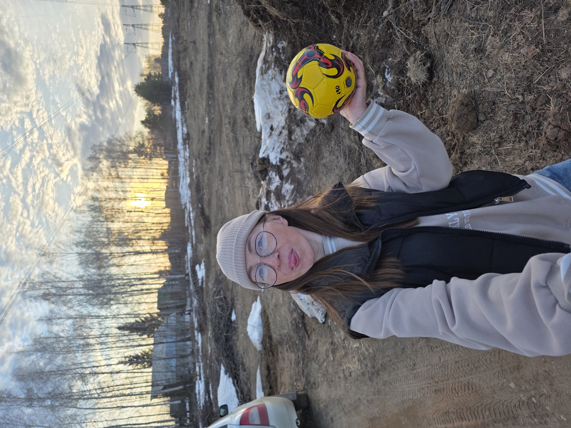 A young woman stands outside in a rural or semi-rural location near Noviy, Russia, holding a yellow soccer ball in her right hand. She wears a beige knit hat, round-framed glasses, a light beige sweatshirt, and a black vest. She is sticking her tongue out. The ground is dirt with patches of snow. In the background, there are trees, power lines, and a sunset. A car is partially visible in the foreground.