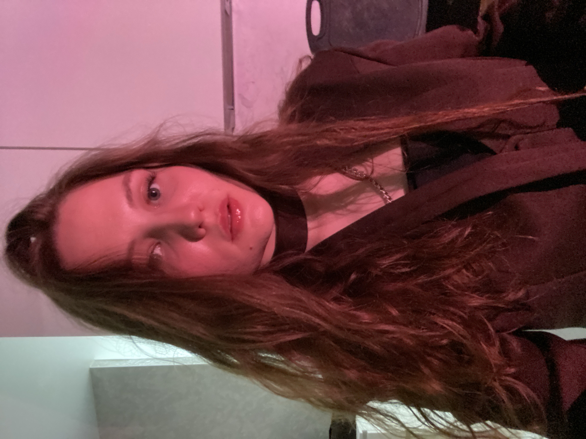 A woman with long, wavy, brown hair is facing the camera. She is wearing a black choker and a dark jacket. There is a silver chain around her neck. The lighting is pink. The location is unknown and cannot be determined from the image.