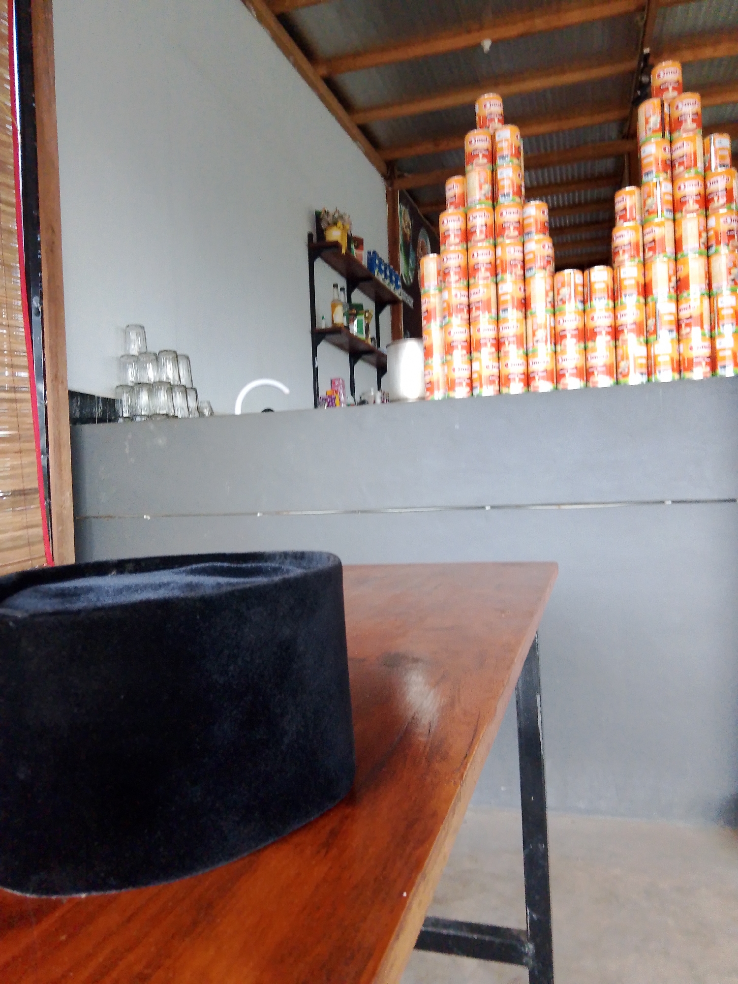 A black peci (a traditional cap) sits on a wooden table in the foreground. In the background, stacks of cylindrical orange cans are arranged on shelves. To the left, a stack of clear glasses is visible, and further left, a bamboo blind hangs on a wall. The scene appears to be indoors, possibly a shop or a storage area in Keudejungkagajah, Indonesia.