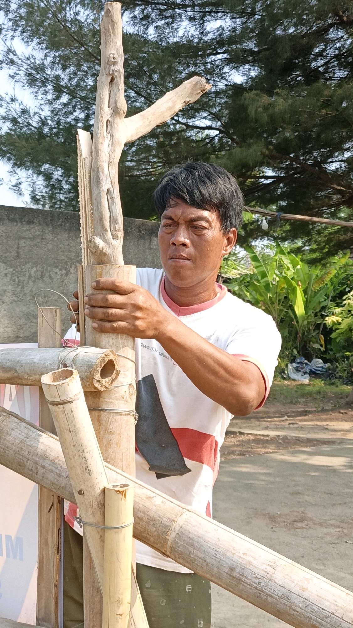 A man, appearing to be of Indonesian descent with dark hair, is constructing something out of bamboo and wood outdoors. He is wearing a white t-shirt with red and black accents and green patterned pants. He is holding a piece of wood and working with bamboo poles that are bound together with wire. The background shows a gray concrete wall, lush green foliage, and trees, suggesting a tropical or subtropical environment. The ground is dirt or gravel. The scene is set in the city of Cikal, Indonesia.