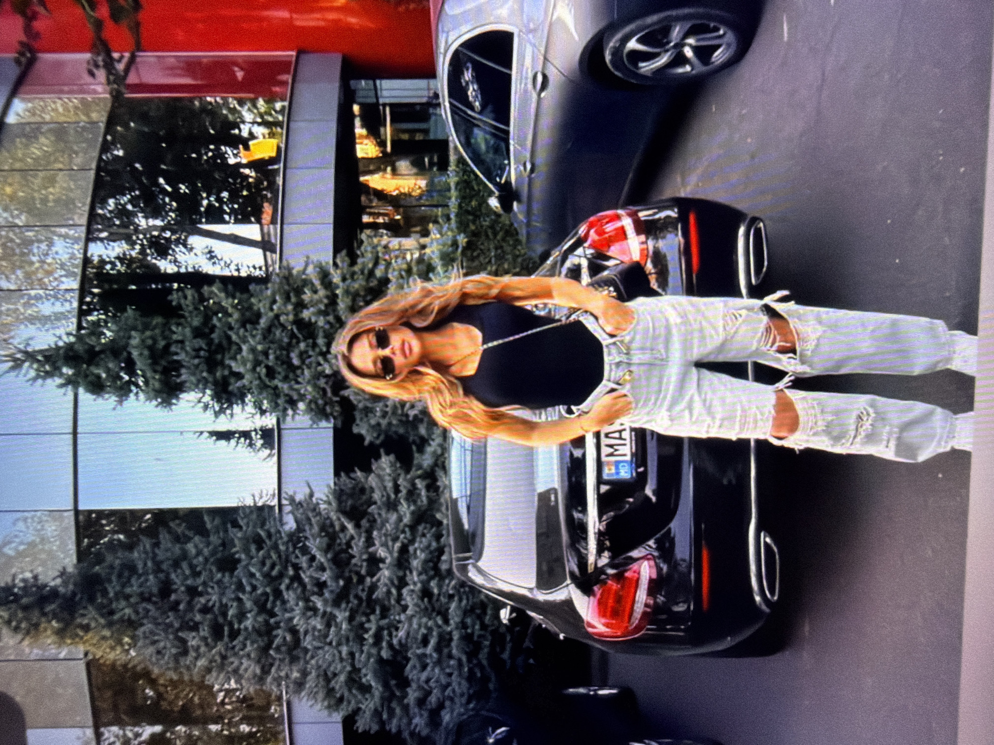 A woman stands next to a black car, her body angled slightly away from the camera. She has long, blonde hair and is wearing sunglasses, a black tank top, and ripped, light-colored jeans. The car has a license plate that reads "MA". The scene appears to be outdoors, with a modern building featuring large glass windows and a red accent visible in the background. Evergreen trees are also present, suggesting a possibly temperate climate. The lighting suggests it might be daytime, possibly late afternoon given the shadows. The woman's stance and expression are neutral, and the overall atmosphere seems casual. The image is a photograph, with a slightly grainy quality.