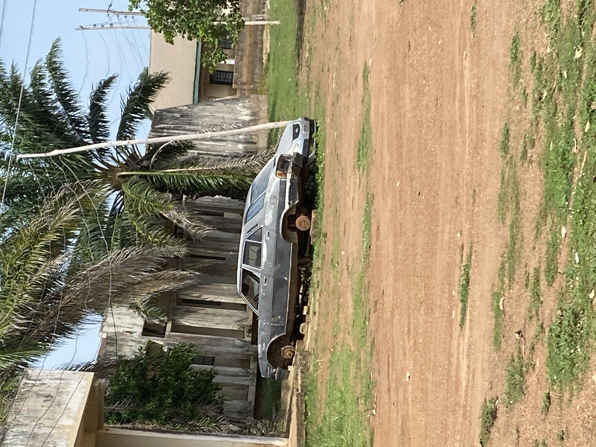 In an outdoor environment, two abandoned cars are parked on a patch of grass next to a dirt road. The car in the foreground, a dark sedan, appears to be missing all of its wheels and is resting on its frame. The car behind it, a lighter-colored sedan, is partially obscured. In the background, a weathered concrete building with palm trees in front of it suggests an abandoned or neglected area, possibly in a tropical or subtropical climate. The sky is clear and blue. The overall scene depicts a state of disrepair and abandonment.