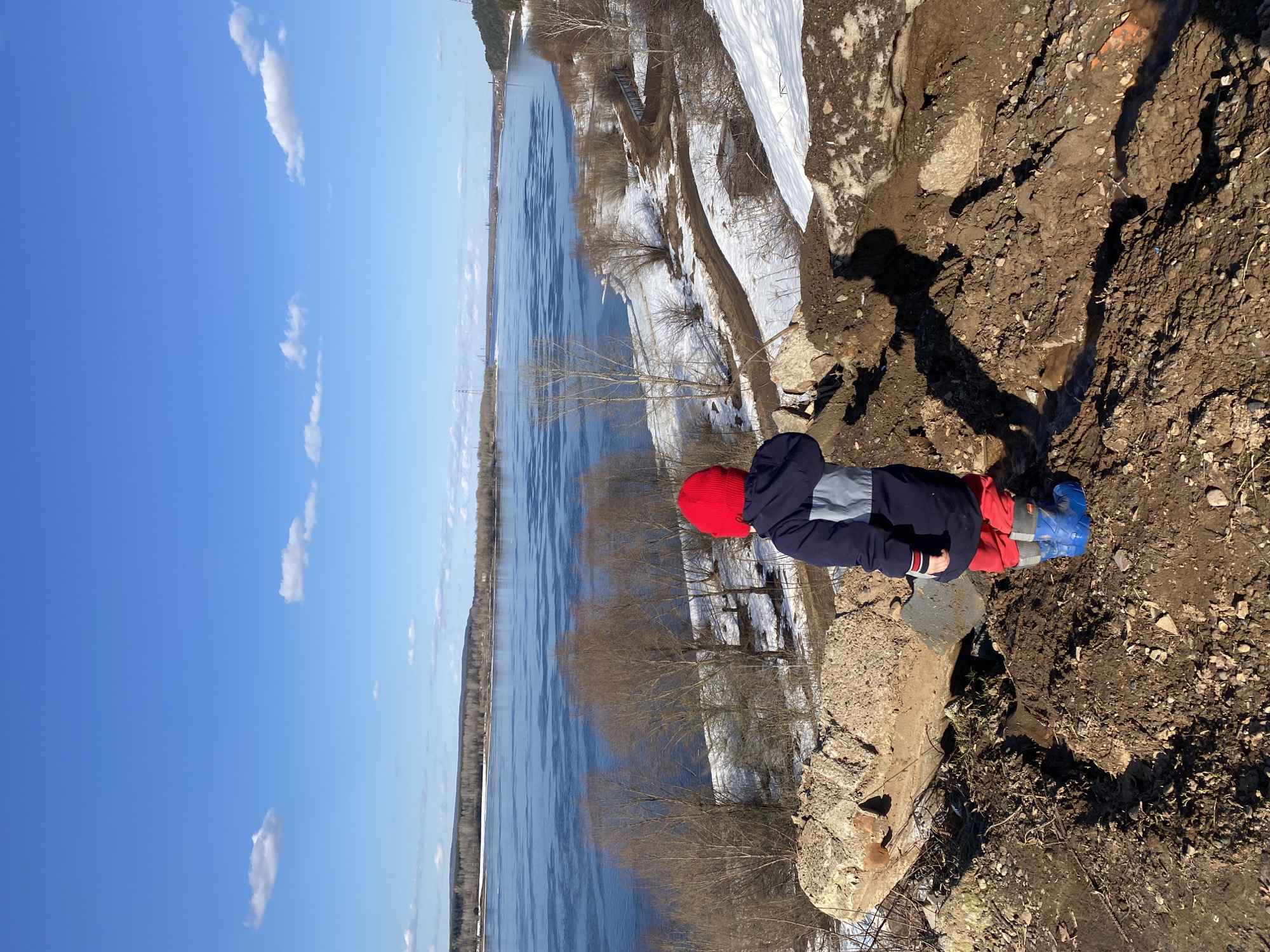 A horizontal outdoor shot shows a child near a riverbank in Ольховка, Russia. The child, wearing a red hat, blue and gray jacket, red pants, and blue boots, is situated on a concrete structure at the edge of a dirt embankment. The river is partially frozen, with trees lining the banks. Patches of snow are visible on the opposite bank. The sky is blue with scattered clouds. The terrain is a mix of dirt and grass.