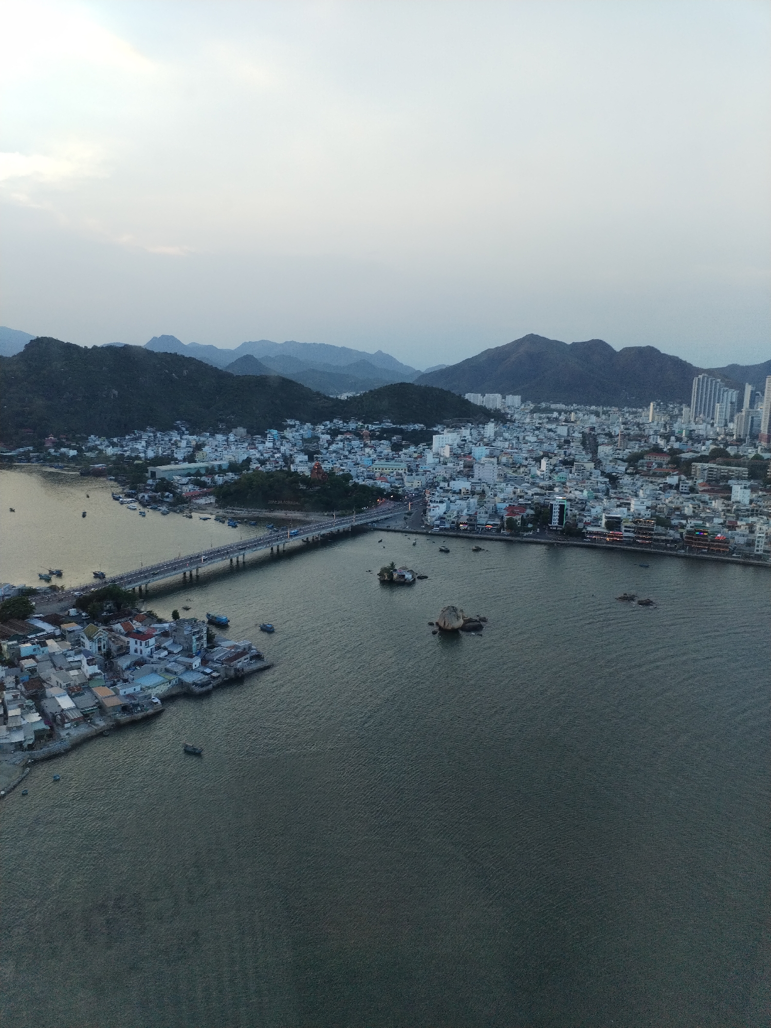 The image is an aerial view of Nha Trang Ward, Vietnam. Mountains are in the background, with a cityscape featuring a mix of low-rise and high-rise buildings closer to the viewer. A bridge spans a body of water, connecting the main part of the city to a smaller cluster of buildings. Several boats are on the water, and a few small islands dot the bay. The sky appears overcast.