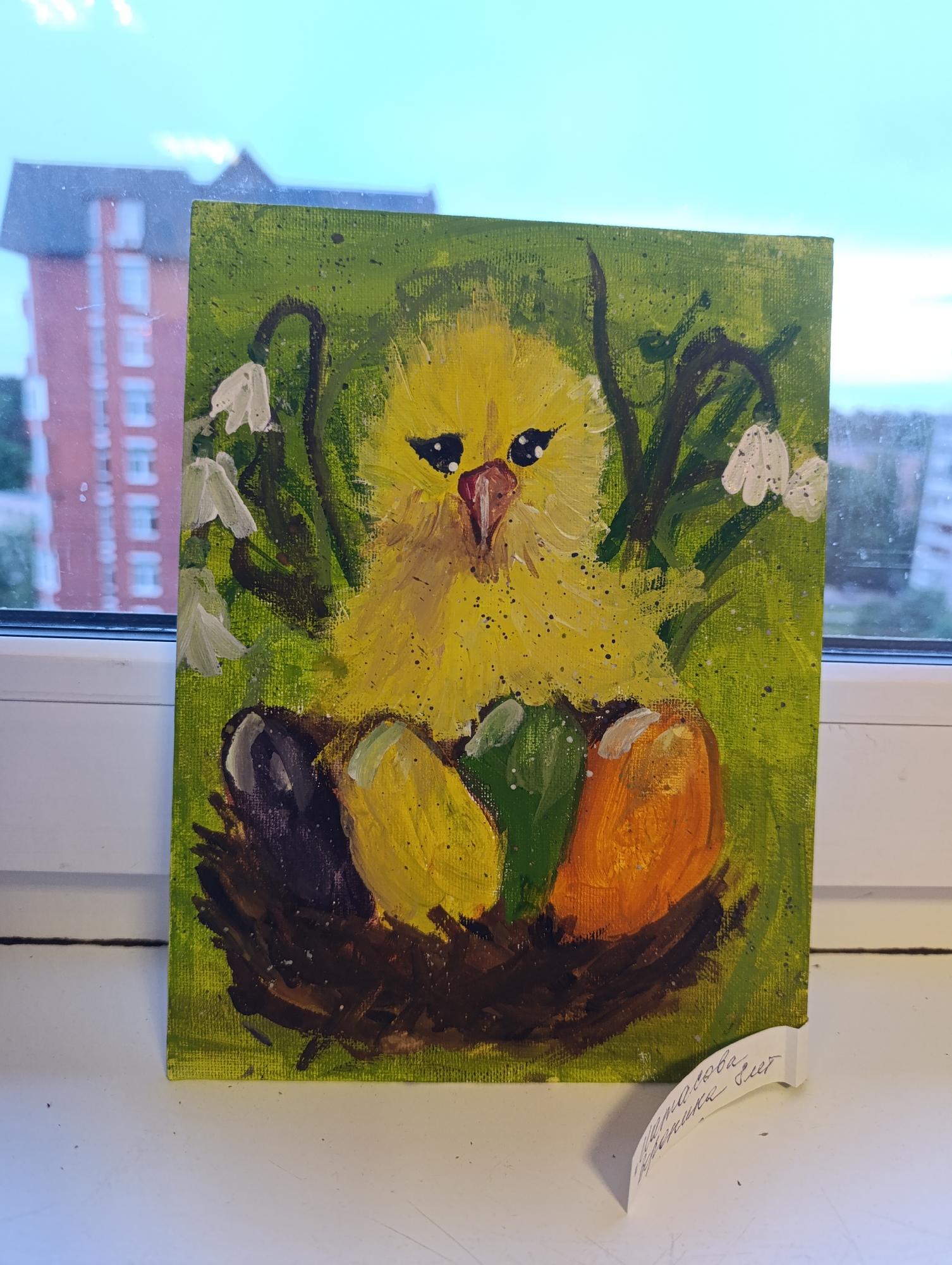 A painting of a yellow chick in a nest of eggs is on display, likely in a private home setting, as it rests on a window sill. The background shows a blurred urban landscape with a multi-story brick building visible through the window. The painting is vibrant with a green backdrop and white flowers painted around the chick. The nest contains eggs colored black, yellow, green, and orange. A handwritten label, seemingly a title card for the painting, is attached to the bottom corner.