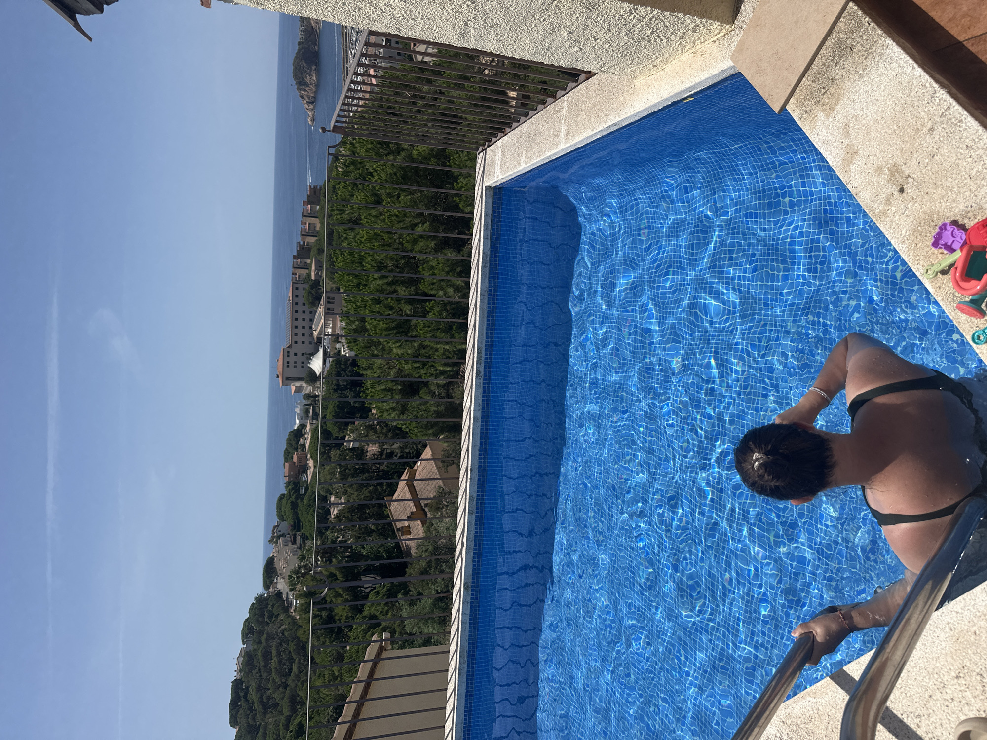 The image depicts a person in a swimming pool with a scenic view of the sea and a coastal town. The pool is filled with bright blue water, tiled with small blue mosaic squares. A person, viewed from behind, is partially submerged in the water, holding onto the pool ladder. They are wearing a black bikini top. The setting is a sunny day, indicated by the bright sunlight and clear blue sky.
Beyond the pool, a metal railing separates the pool area from a hillside covered in lush green trees. Scattered amongst the trees are buildings, suggesting a residential or resort area. Further in the distance, the azure expanse of the sea meets the horizon. A small island or rocky outcrop is visible in the sea. The architecture of the buildings in the town appears to be Mediterranean in style, with terracotta roofs.
On the edge of the pool, to the right, are some colorful plastic children's toys, including a purple sand shovel and a red bucket. The overall mood of the image is one of leisure and relaxation, characteristic of a vacation or a sunny day spent by the water. The lighting suggests it is daytime, likely late morning or afternoon. The location is identified as Sant Feliu de Guíxols, Spain, which aligns with the coastal scenery and Mediterranean architecture. There is no visible text in the image.