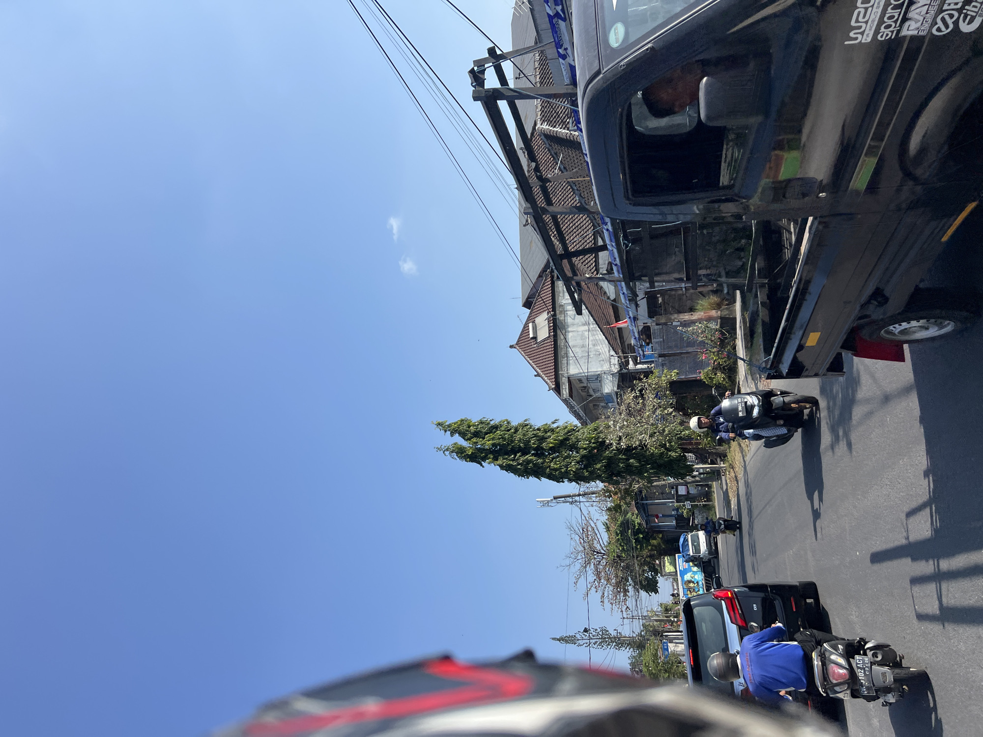 The image depicts a street scene in Denpasar, Indonesia, on a clear, sunny day with a bright blue sky. The perspective is from a vehicle, likely a motorcycle, looking forward. In the foreground, the front portion of a black pickup truck is visible, with a person's head and shoulders seen through the driver's side window. The truck has a metal rack structure in its bed, with blue and white straps. Various decals are present on the rear fender of the truck.
On the road, several other vehicles are present. A black SUV is in the left lane, with a person wearing a blue shirt and a helmet seated on a scooter behind it. Further down the road, a person on a scooter is visible in the right lane, wearing a white helmet. In the distance, another scooter and a small white truck are also on the road.
The environment includes buildings with tiled roofs, some appearing to be shops or residences, lining the street. Tall, slender trees are interspersed with the buildings. Utility poles and electrical wires crisscross the scene. The road surface is asphalt. The overall impression is of a typical urban street in Denpasar during daylight hours, with moderate traffic.