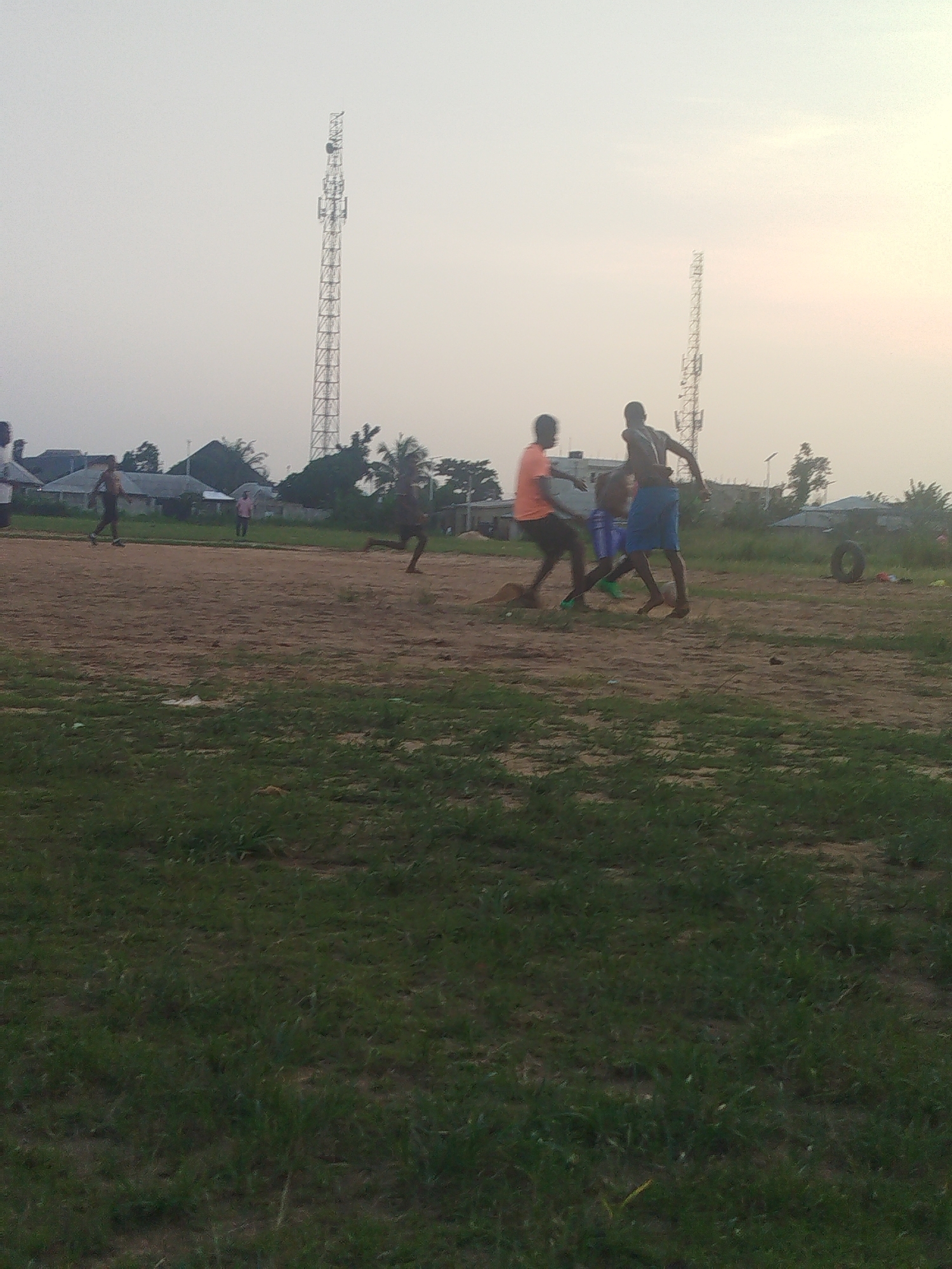 The image shows a group of people, possibly playing a sport like soccer, on a dirt field with patches of green grass. Several individuals are in motion, with at least two appearing to be in pursuit of a ball. Behind them are houses and trees, and in the background, two tall communication towers are visible. The setting appears to be outdoors during daylight hours, likely in the vicinity of Ikot Mbonde, Nigeria.