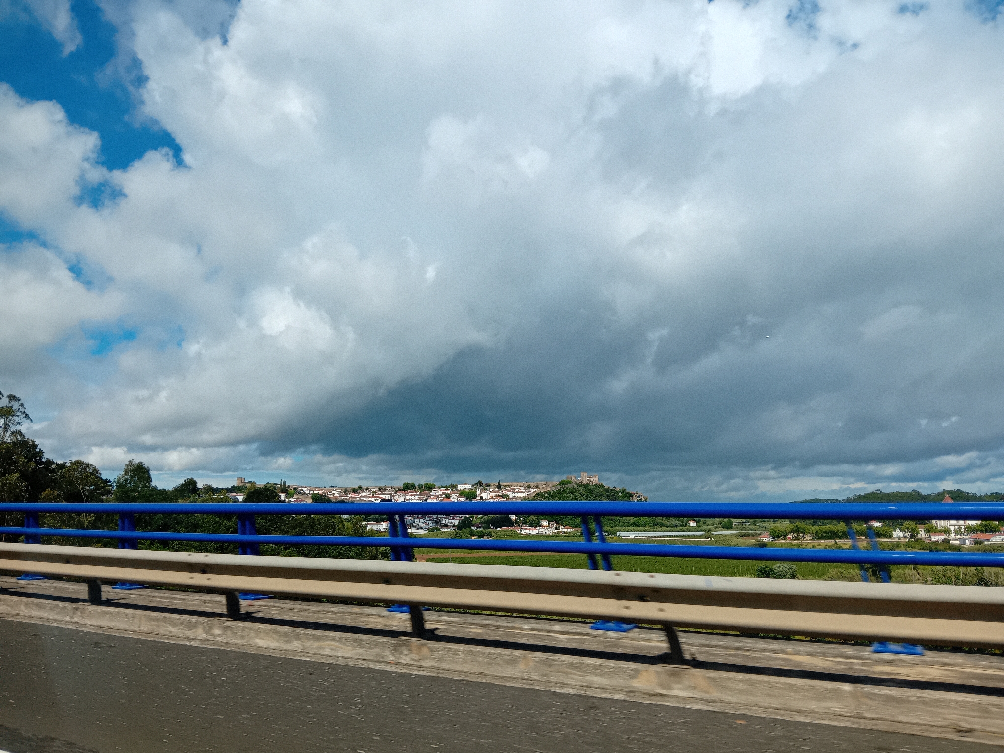 The image depicts a view from a bridge or elevated road, overlooking a landscape that includes a town or city. The foreground features the bridge's infrastructure, specifically the guardrails, which are colored blue and beige. Beyond the guardrails, there is a view of greenery, possibly trees or shrubs, and a field. In the distance, there is a town or city with white buildings and a castle visible atop a hill. The sky is dominated by thick, grey clouds, with a patch of blue sky visible in the upper-left corner. The road surface is asphalt.