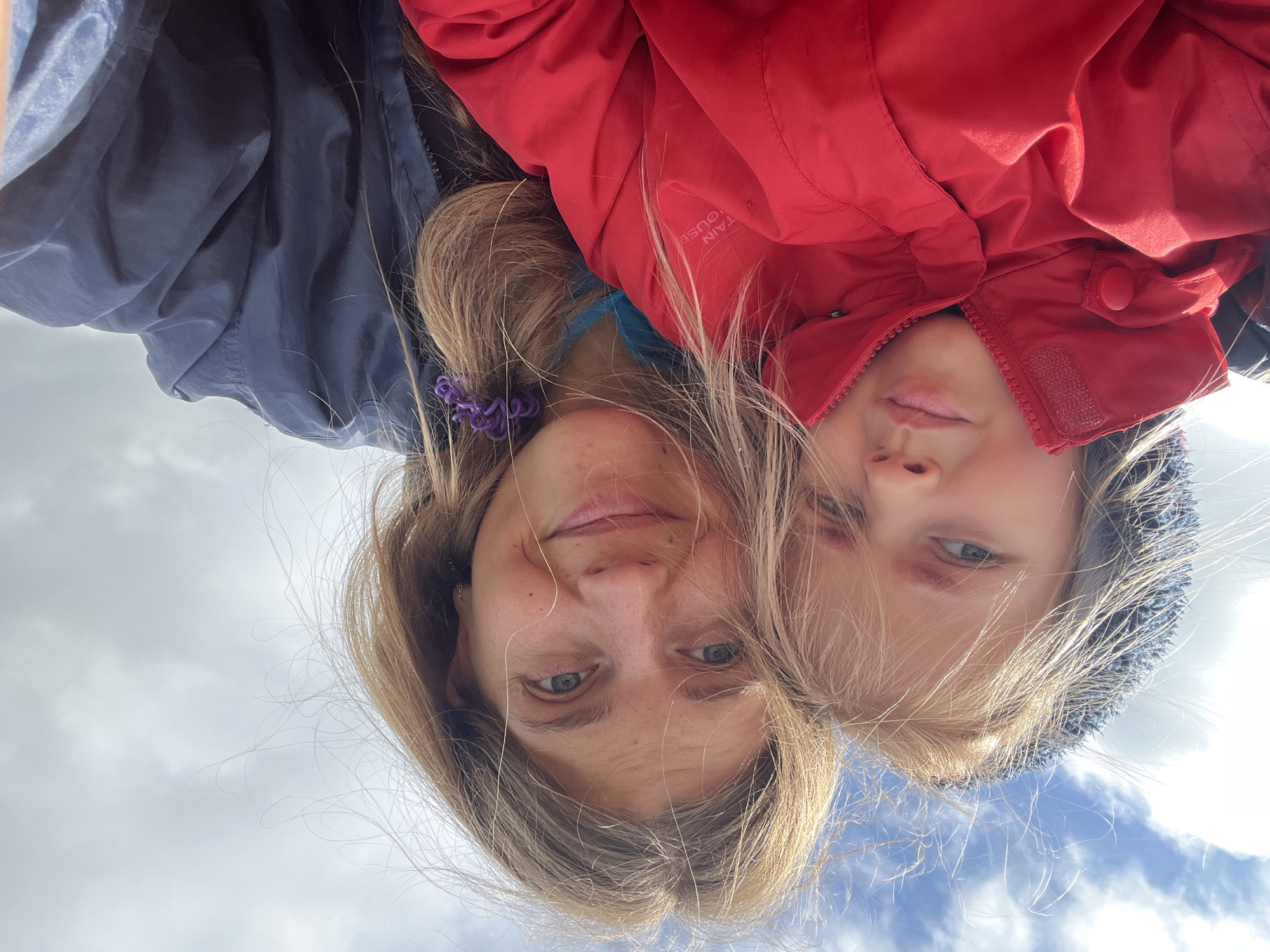 Two individuals, a child and an adult, are captured in a close-up, selfie-style photograph outdoors under a partly cloudy sky. The child, positioned on the left, wears a red jacket with the hood up, partially obscuring their head. They have blonde hair that is being blown by the wind. Their gaze is directed towards the camera. The adult, on the right, is looking at the camera and smiling. They have blonde hair that is also windswept and is styled with a purple hair tie. They are wearing a dark blue jacket over a blue garment. The background consists of a sky with clouds, suggesting an open outdoor environment, possibly a park or natural area. The image appears to be taken during daylight. There is no discernible urban environment present; therefore, the location is not within the city of Дунілавіцкі сельскі Савет, Belarus, as there are no identifying landmarks.