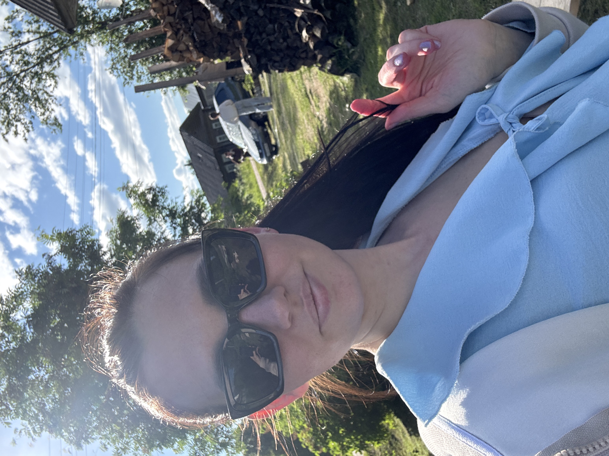 The image is a selfie taken by a young woman outdoors, rotated to a vertical orientation in the file but depicting a horizontal scene. The woman is the primary subject, positioned in the foreground and looking directly at the camera with a relaxed expression. She has long, dark hair, visibly illuminated by strong sunlight, and is wearing dark, rectangular sunglasses. Her attire includes a light blue top with a ruffled or layered neckline, possibly tied at the chest, worn over a white undershirt. Her left hand, adorned with light, shimmery nail polish, is gently touching a strand of her hair.
The background suggests a rural or semi-rural setting in Луптюг, Russia. The sky is bright blue with scattered white clouds, indicating clear and sunny weather during daytime. Green foliage from trees frames the scene and extends into the mid-ground. Visible in the distance are several structures: a dark wooden house or building with multiple windows, a light-colored car (possibly white or light blue) parked near the building, and a large woodpile stacked beneath an overhang or shed. Green grass covers the ground in the background. Thin, dark vertical lines, possibly power lines, are faintly visible against the sky. The strong sunlight creates a luminous effect on the woman's hair and clothing. No text is visible in the image.