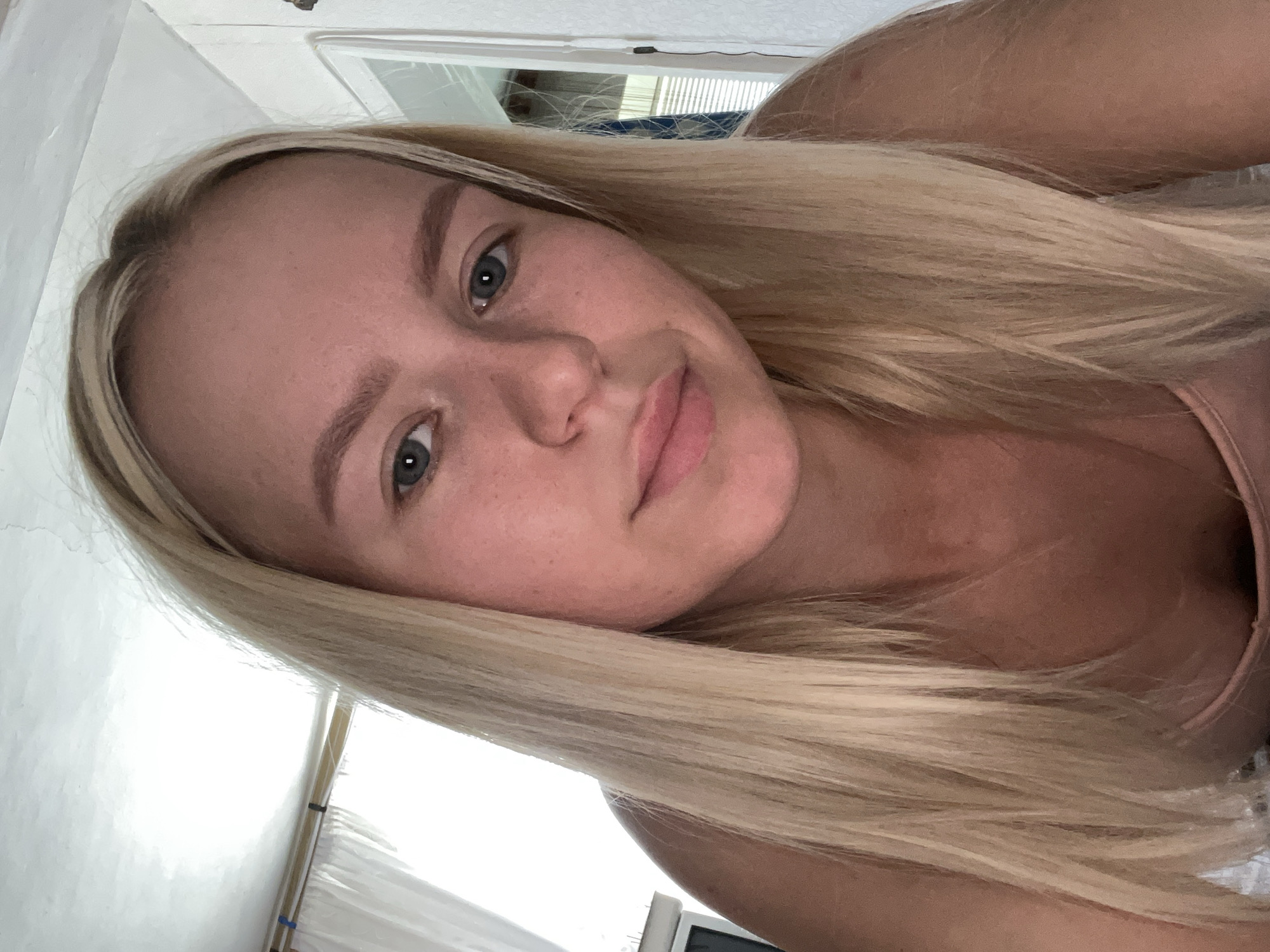 A young woman with blonde hair is looking at the camera. She is wearing a pink top. The background appears to be a room with white walls and a window with white curtains. The lighting is bright and natural.
