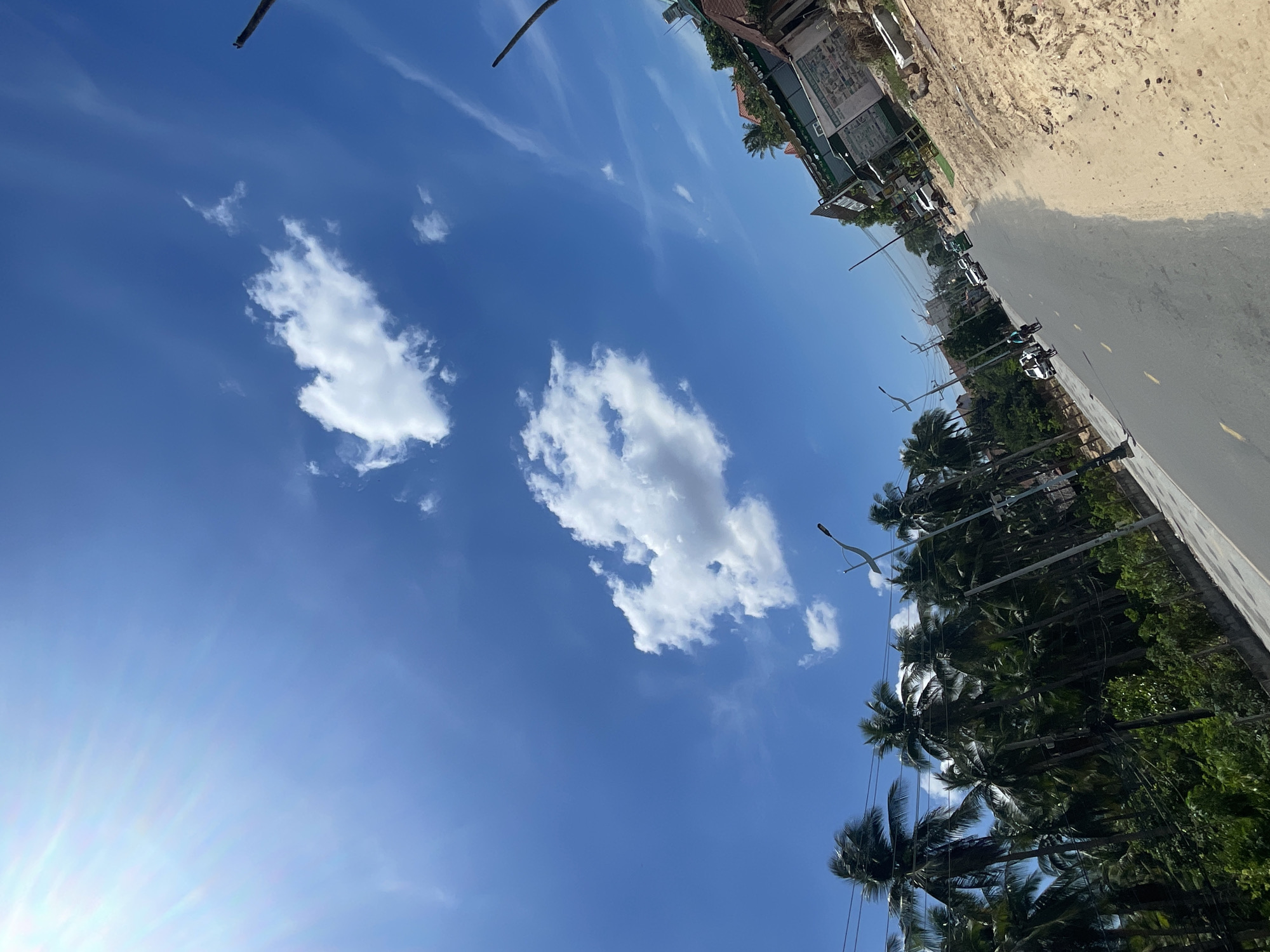 The image captures a sun-drenched street scene in Phu Thuy Ward, Vietnam. A bright blue sky with scattered white clouds dominates the upper portion of the frame, with sun rays emanating from the top left. The foreground features a paved road with yellow lane markings, sloping towards the right. On the left side of the road, a dense line of tall palm trees with lush green foliage stretches into the distance. A few utility poles and streetlights are visible along this side.
On the right side of the road, a construction site is evident, with piles of sand and dirt. Several buildings are present, including one with a green facade and what appears to be a sign with text and images. Further down the road, a few vehicles, including cars and a bicycle with a rider, are moving. The overall impression is one of a sunny day in a tropical or sub-tropical urban environment, possibly on the outskirts of a city.