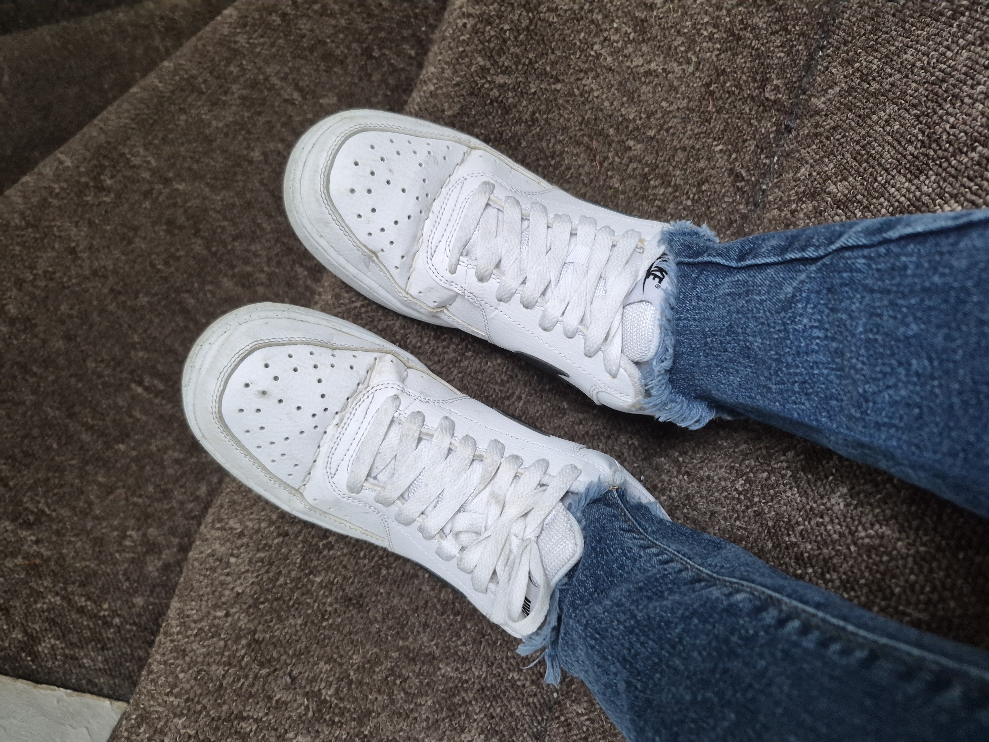 A person is sitting on a brown carpeted staircase, wearing distressed denim jeans and white Nike sneakers. The jeans have a frayed hemline, and the sneakers appear worn, with scuff marks and small perforations on the toe box. The image is a close-up shot focusing on the lower legs and feet of the individual. The location is identified as a residential property in the city of Sandwell, United Kingdom.