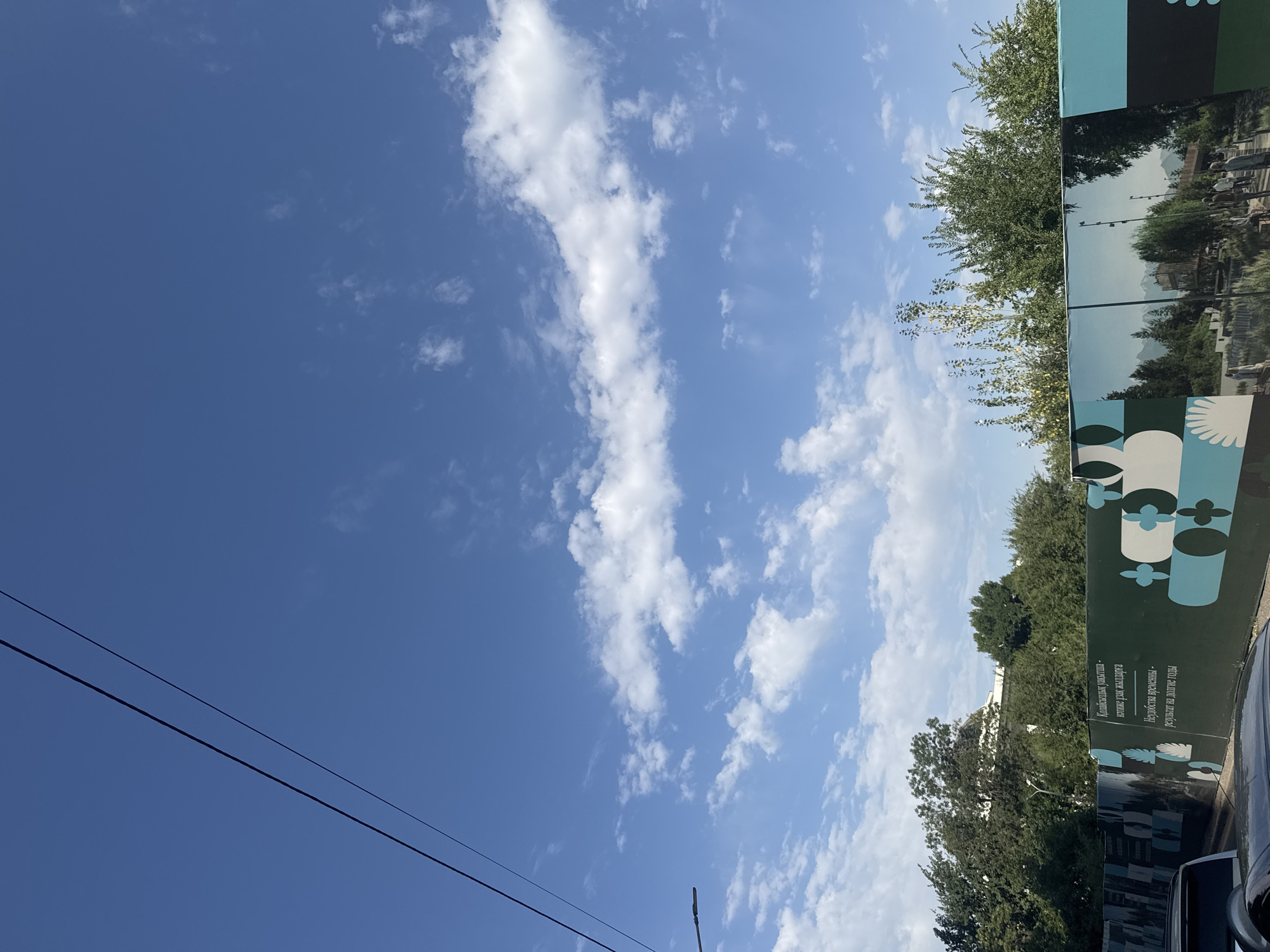 The image shows a clear blue sky with scattered white clouds. Two power lines stretch diagonally across the upper portion of the frame. Below the sky, a dense line of green trees forms the horizon. In the foreground, a construction fence is visible, adorned with a large graphic mural. The mural depicts geometric patterns in teal and white on the left side, and a scenic landscape with people walking on the right. The landscape features mountains in the background, pathways, trees, and an architectural structure. The bottom of the image shows parts of two cars, suggesting an urban setting. The mural's text, visible on the fence, is in Kazakh and Russian and appears to be a slogan related to development or progress. Given the architecture and the Russian text on the mural, this scene is likely in Almaty, Kazakhstan, possibly in a park or public space undergoing renovation.