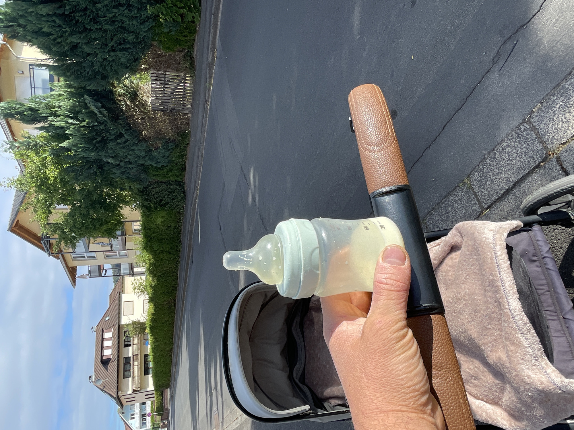 A person's hand holds a baby bottle filled with milk next to a stroller on a street in Neukirchen, Germany. The stroller has a brown leather handle and a pink blanket inside. In the background, there are houses and trees. The sky is cloudy.