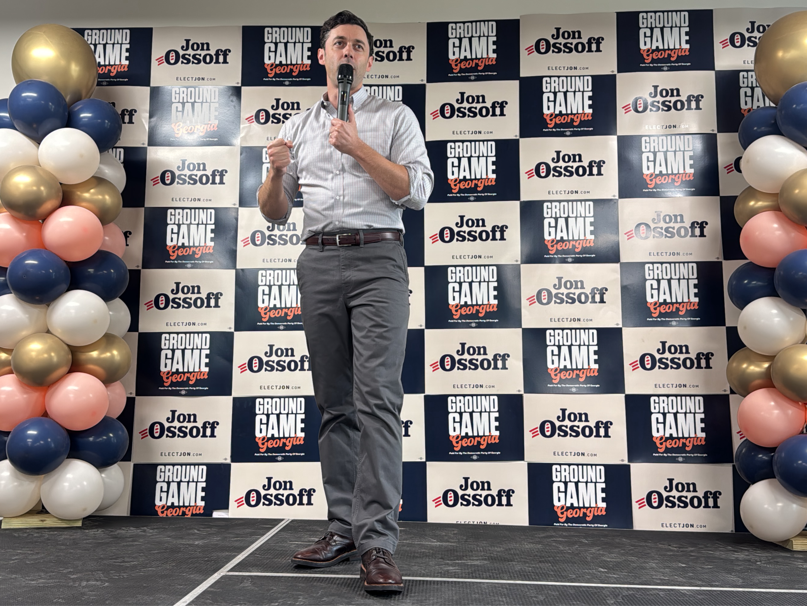 The image shows a man, presumably Jon Ossoff, standing on a stage with a microphone in his hand, addressing an audience. He is wearing a light-colored button-down shirt, gray pants, and brown leather shoes with a belt. Behind him is a backdrop with a repeating pattern of "GROUND GAME Georgia" and "Jon Ossoff ELECTJON.COM" on alternating black and white squares. To his left and right are balloon arrangements consisting of navy blue, gold, white, and peach-colored balloons.
The setting appears to be an indoor event, possibly a political rally or campaign event. The image was taken in Decatur, United States.