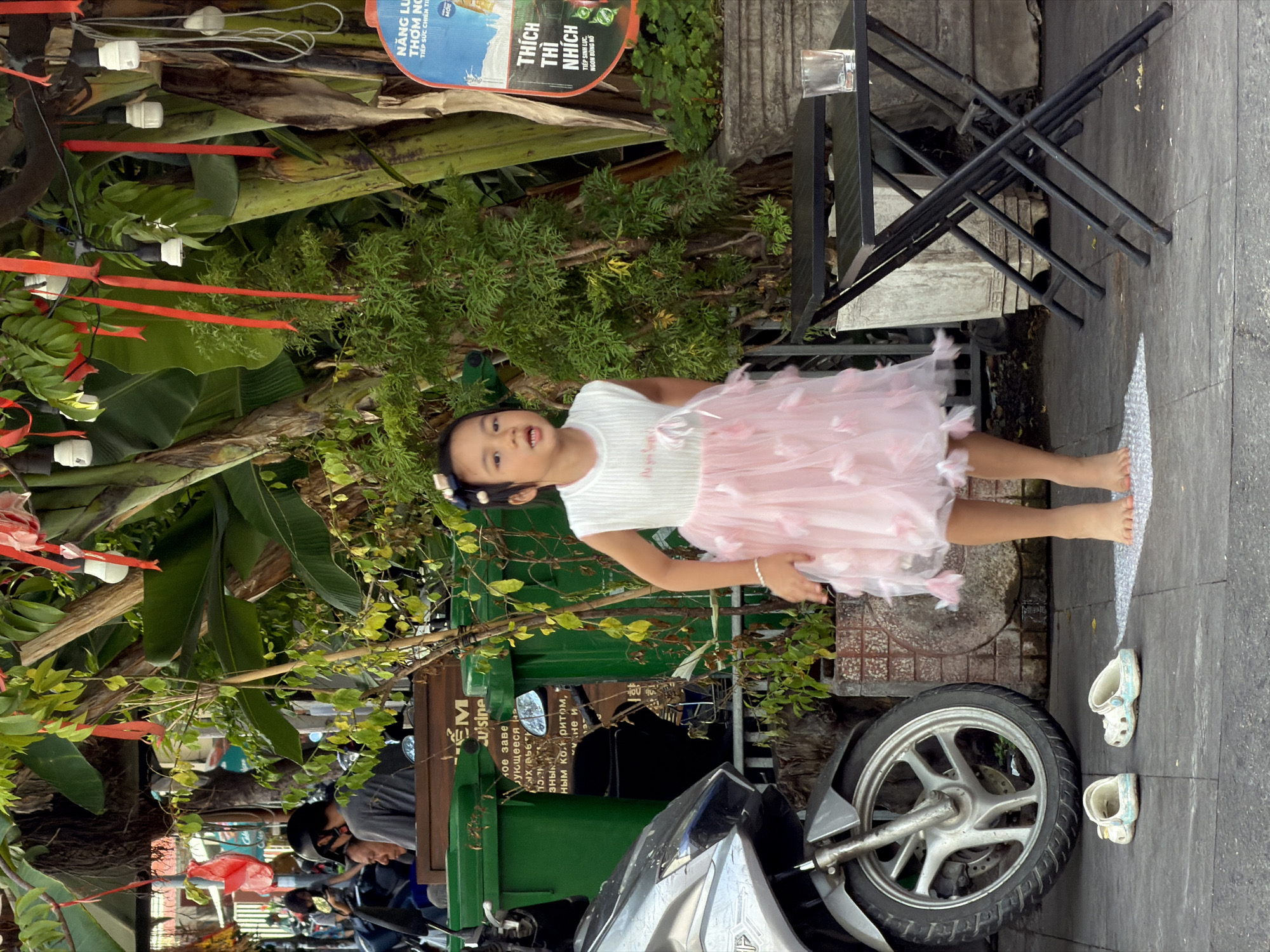 A young girl in a pink tutu and white top stands on a paved walkway next to a motorcycle, her body tilted slightly to the left. She has a slight smile on her face. Her bare feet are visible, as well as a pair of light blue Crocs lying on the pavement. Behind her, lush green foliage, including large banana leaves and smaller bushes, creates a vibrant backdrop. A black metal table and chairs are also visible to the left. A sign with Vietnamese text, including "NĂNG LU THƠM NG" and "THÍCH THÌ NHÍCH," is partially visible. The scene appears to be outdoors, possibly in a cafe or restaurant setting in Nha Trang, Vietnam, during the daytime. The overall mood is casual and playful.