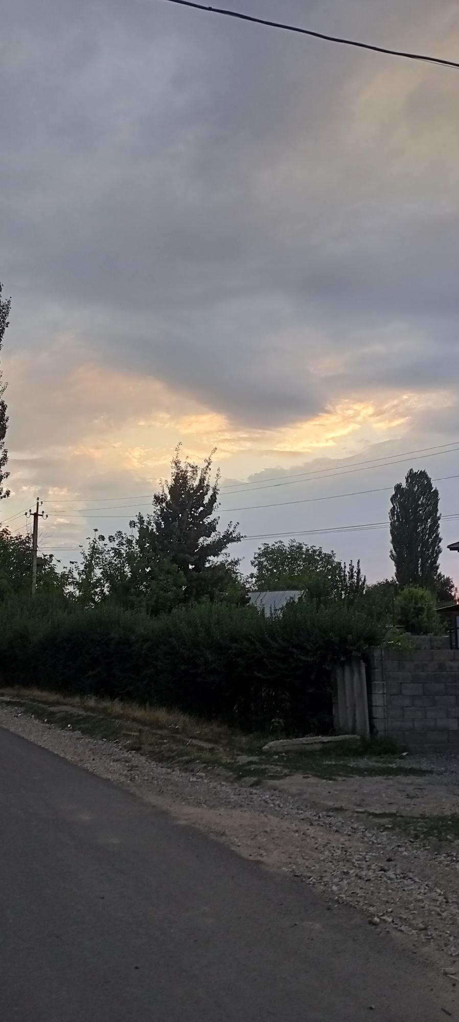 The image depicts a rural roadside scene in Sokuluk, Kyrgyzstan, under a cloudy sky with patches of warm light breaking through. A paved road occupies the foreground, transitioning to a gravel shoulder. To the right, a low brick wall with a concrete pillar borders a property. Beyond the wall and a line of dense green bushes, a corrugated metal roof is visible among trees. A utility pole with wires stands on the left side of the road, and several trees, including a tall, slender one on the right, punctuate the landscape. The overall impression is one of twilight or a dimly lit day in a semi-urban or rural setting.