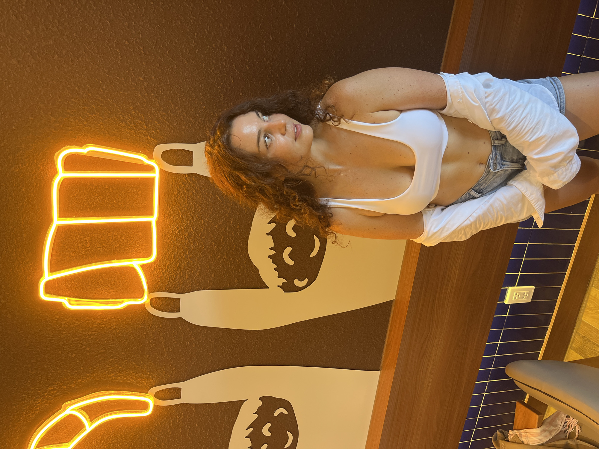 The image captures a woman with curly brown hair wearing a white bikini top and denim shorts, with an open white shirt draped over her arms. She is standing in front of a brown textured wall adorned with neon yellow light art depicting mugs and stylized figures. To her left, the wall features large, light-colored silhouettes of cartoonish faces with happy expressions, each emerging from a long, slender shape. A wooden shelf runs horizontally across the lower part of the wall, with blue tiled accents below it. The scene appears to be an indoor establishment, possibly a bar or cafe, with a portion of a chair visible in the lower-left corner and an electrical outlet on the tiled section of the wall. The lighting is warm and inviting, highlighting the neon artwork and the woman's profile as she looks away from the camera. The overall ambiance suggests a casual and relaxed setting within the city of Phan Thiết, Vietnam.