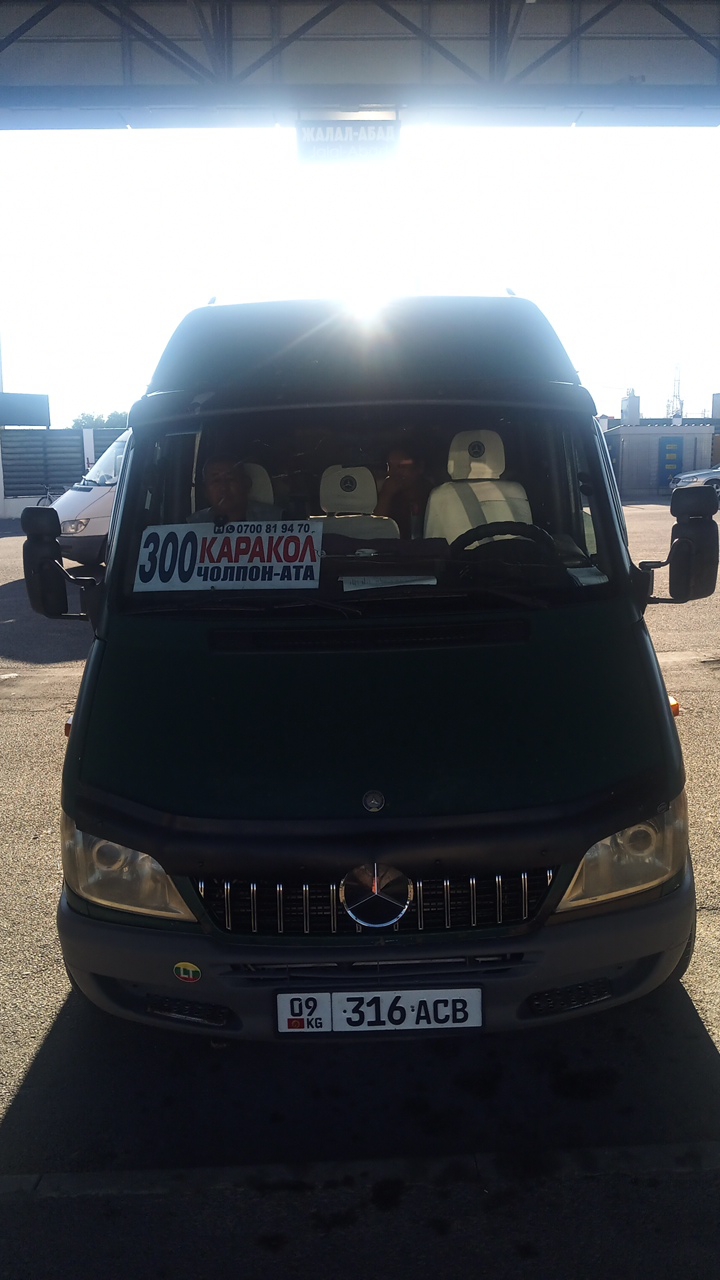 A dark green Mercedes-Benz Sprinter van, identified by the license plate "09 316 ACB", is parked in an outdoor area, possibly a bus station or depot, under a covered structure. A sign above the van reads "ЖАЛАЛ-АБАД", indicating a destination or origin. The van's windshield displays a sign with the text "300 КАРАКОЛ ЧОЛПОН-АТА" and a phone number "0700 81 94 70". Inside the van, at least three passengers are visible, two in the front seats and one further back. The driver, a man, is seated in the driver's seat and appears to be smoking. The van has prominent Mercedes-Benz logos on the grille and headrests. The environment is bright and sunny, suggesting daytime. The van is positioned on an asphalt surface, with shadows cast on the ground. A white van is parked to the left of the Mercedes-Benz.