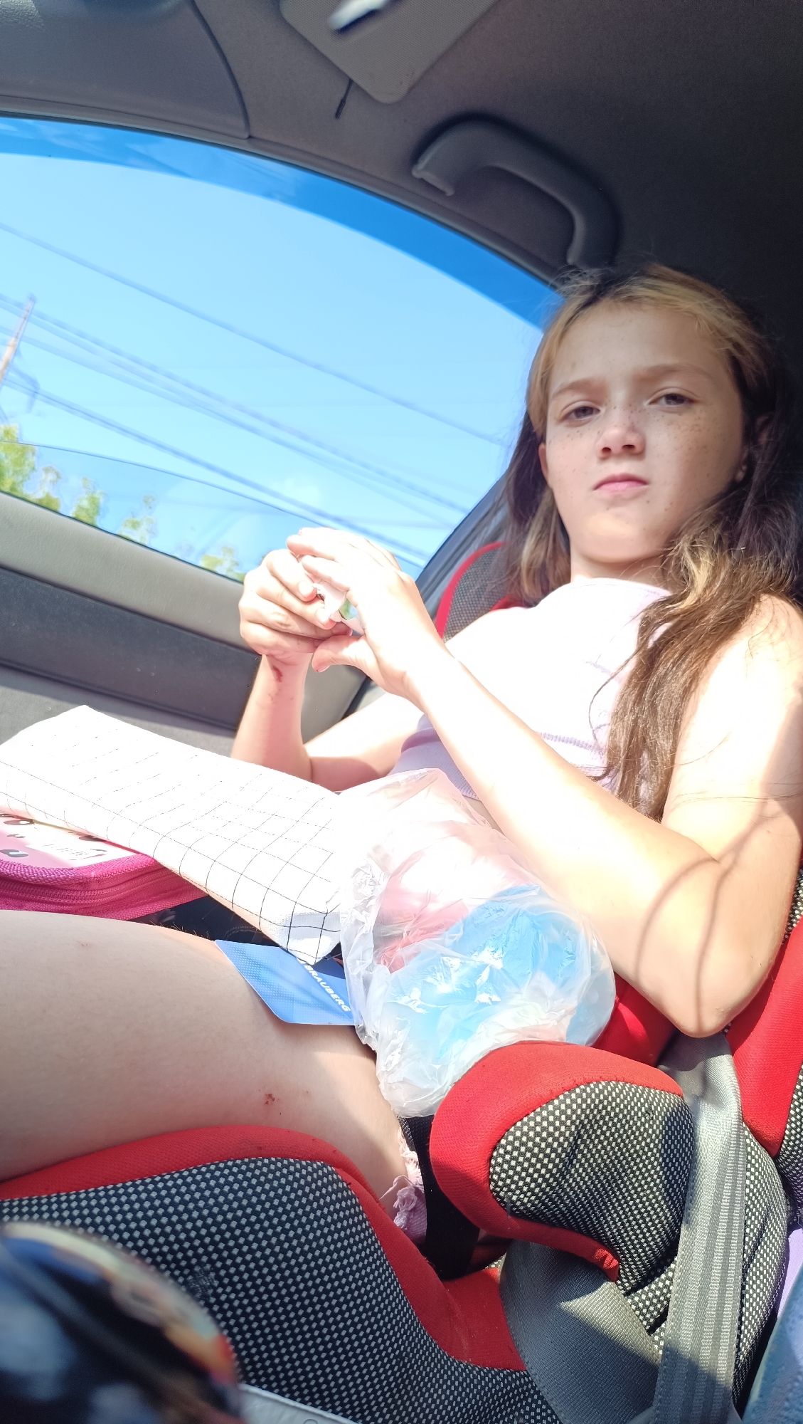 The image shows a young girl sitting in a car seat, looking towards the camera with a neutral expression. She has long, light brown hair with some blonde highlights and freckles on her face. She is wearing a light-colored top. Her hands are clasped together, and she appears to be holding a small white object. Next to her is a notebook with a checkered pattern and a pink cover, and a plastic bag filled with what appear to be blue and possibly red items. A small blue tab with text is visible on the notebook.
The setting is the interior of a car, with the blue sky and some power lines visible through the window. The sunlight suggests it is daytime, likely mid-morning or afternoon. The girl's gaze and the overall composition give a sense of a candid moment, possibly during a car journey. Her posture and expression do not convey any strong emotions. The background is slightly blurred, focusing attention on the girl and the objects in the foreground.