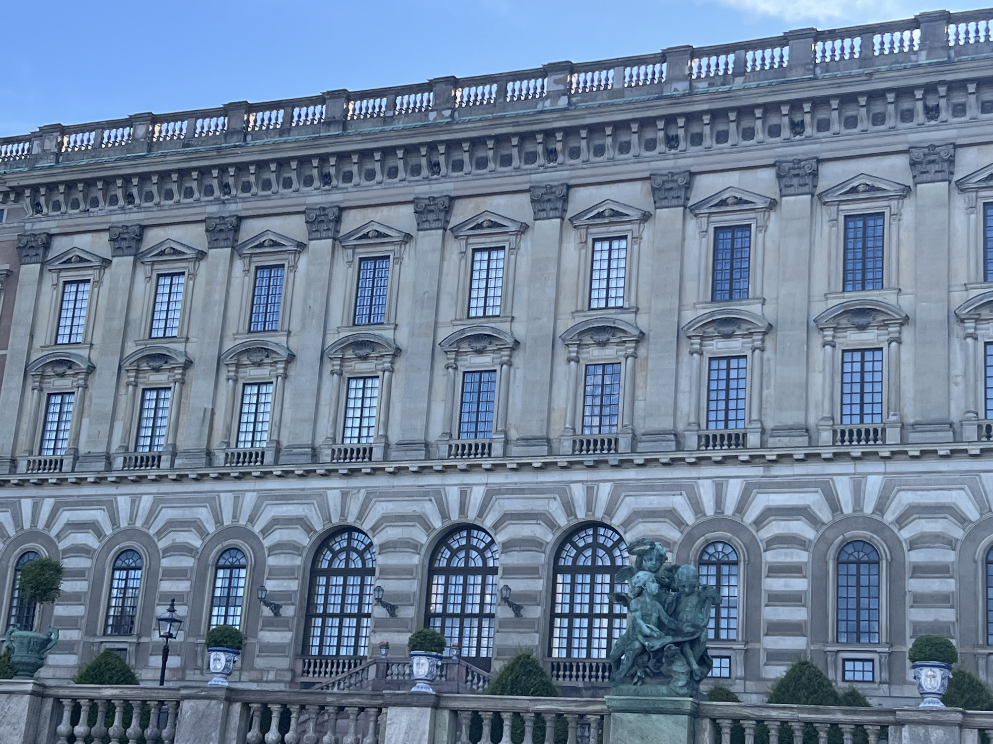 The image shows the Royal Palace in Stockholm, Sweden, captured from a slightly low angle, emphasizing its grand architecture. The sky above is a clear blue, with a hint of clouds. The palace facade is constructed of light-colored stone and features multiple stories with numerous windows. The windows on the upper floors are rectangular with dark frames, arranged in rows and separated by pilasters. The lower floors have arched windows with decorative surrounds. A balustrade runs along the top of the building, and elaborate cornices and decorative elements are visible throughout. In the foreground, there is a stone balustrade, and to the right of the image, a bronze sculpture depicting several cherubic figures is positioned on a stone pedestal. Various potted plants and trees are placed along the balustrade. The overall impression is one of a majestic and historic building.