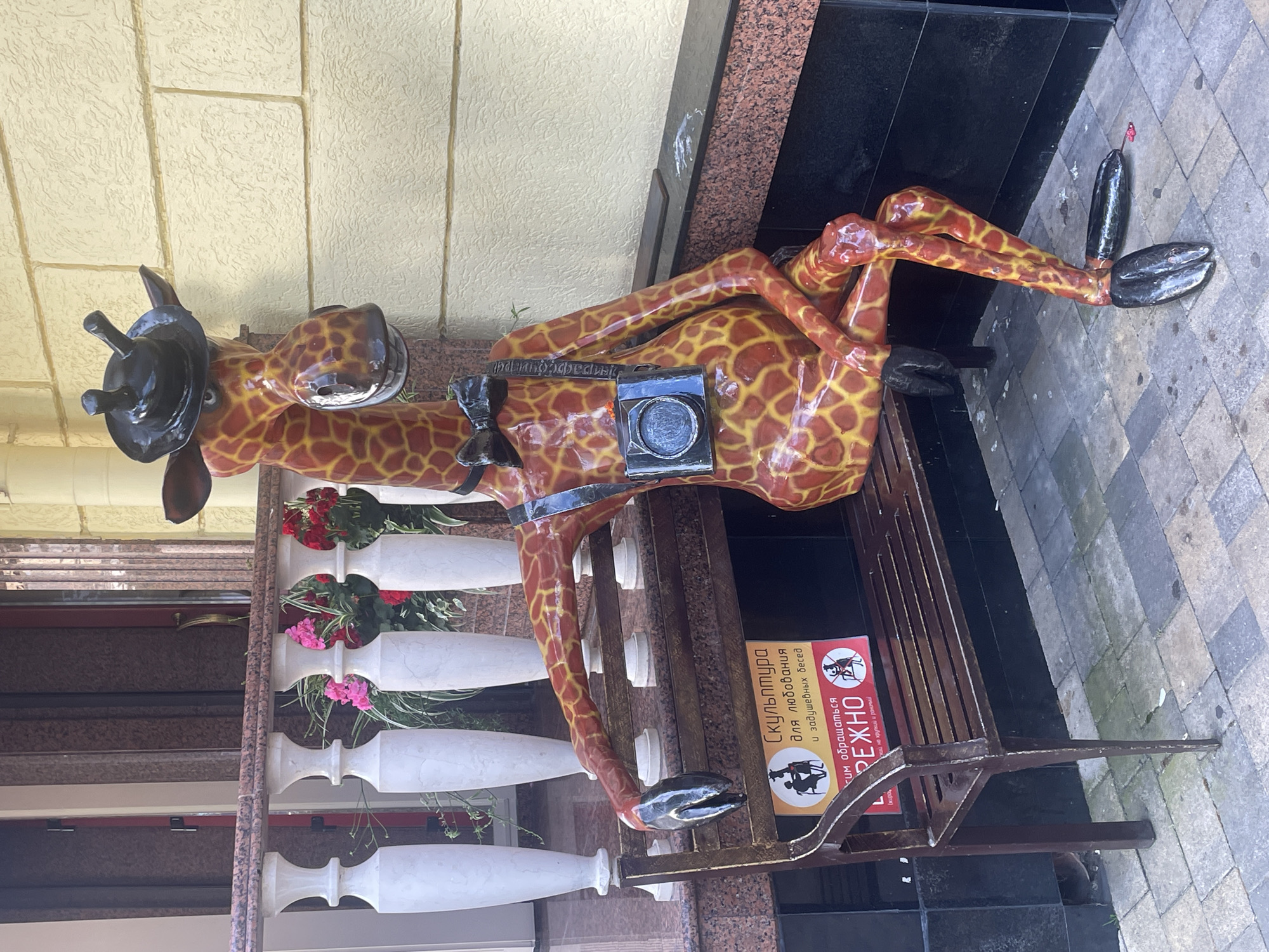 A statue of a giraffe wearing a black hat and a camera around its neck sits on a park bench. The giraffe has an orange and yellow patterned body and black hooves. A sign in Russian on the bench reads "Sculpture for enjoyment and soulful conversations." The background shows a building with light yellow textured walls and decorative white balusters. Red and pink flowers are visible in planters. The scene is outdoors, on a paved area, suggesting a public space. The statue is located in Sochi, Russia.