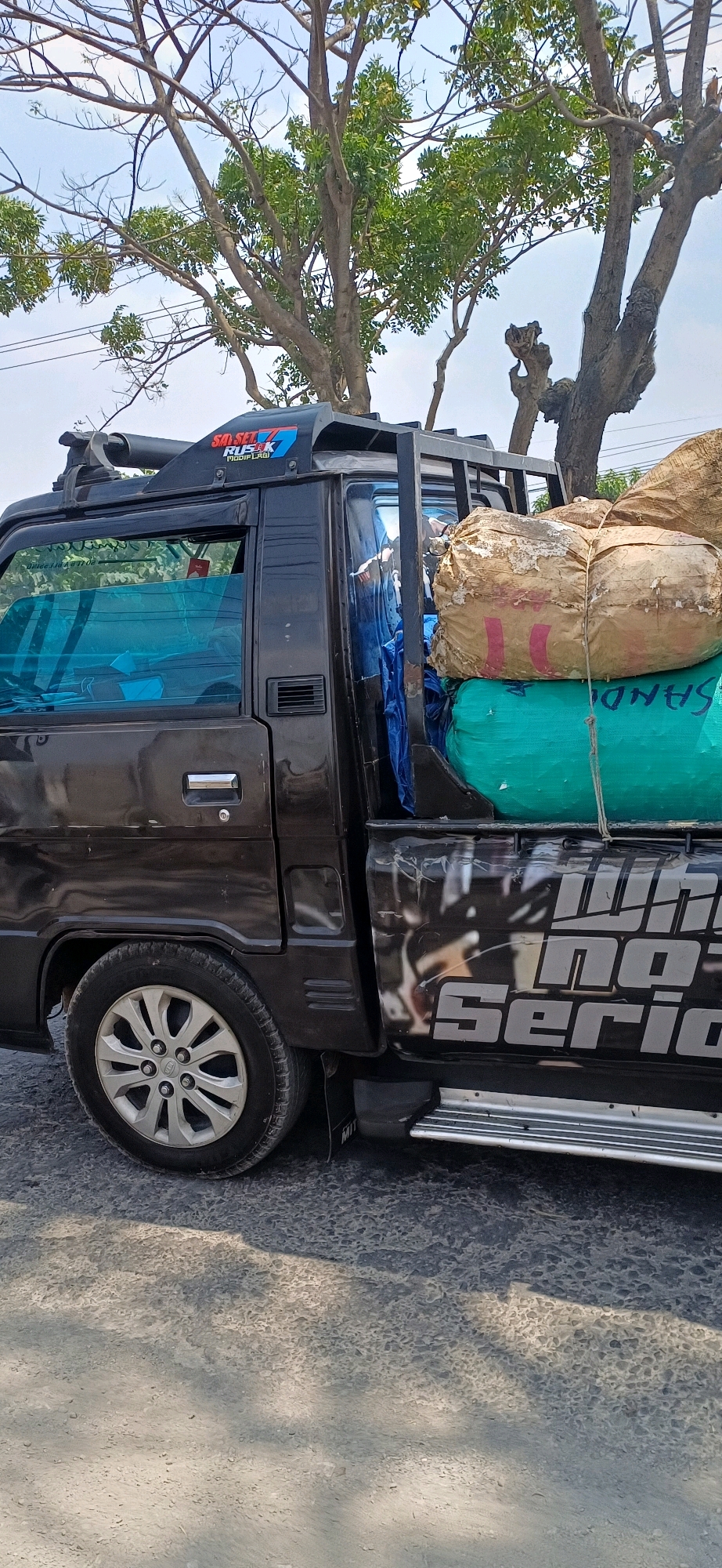 A black pickup truck is parked on a grey, asphalt road, possibly in Kecamatan Pati, Indonesia, with trees and power lines in the background. The truck's bed is loaded with two large sacks of goods, one light brown and tan, and the other green. The truck has tinted windows and a silver roof rack. A chrome step bar runs along the bottom of the truck's passenger side. On the side of the truck, a sticker reads "SO SET RUIST MODIFLAG".