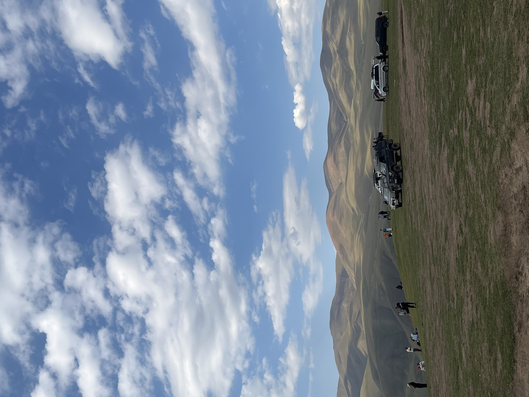 An outdoor landscape image taken in Batan, Kazakhstan, features rolling, grassy hills under a bright blue sky filled with white, scattered clouds. In the foreground, a grassy plateau is dotted with several off-road vehicles, including a dark gray SUV, a silver van, and a black SUV. A group of people, numbering approximately 15 to 20, are scattered across the grassy area, some standing, some sitting, and some appearing to be in conversation. The mid-ground is characterized by more rolling hills, while the background consists of layered, mountainous terrain with patches of reddish soil visible on some slopes. The overall impression is of a scenic natural environment with recreational activity.