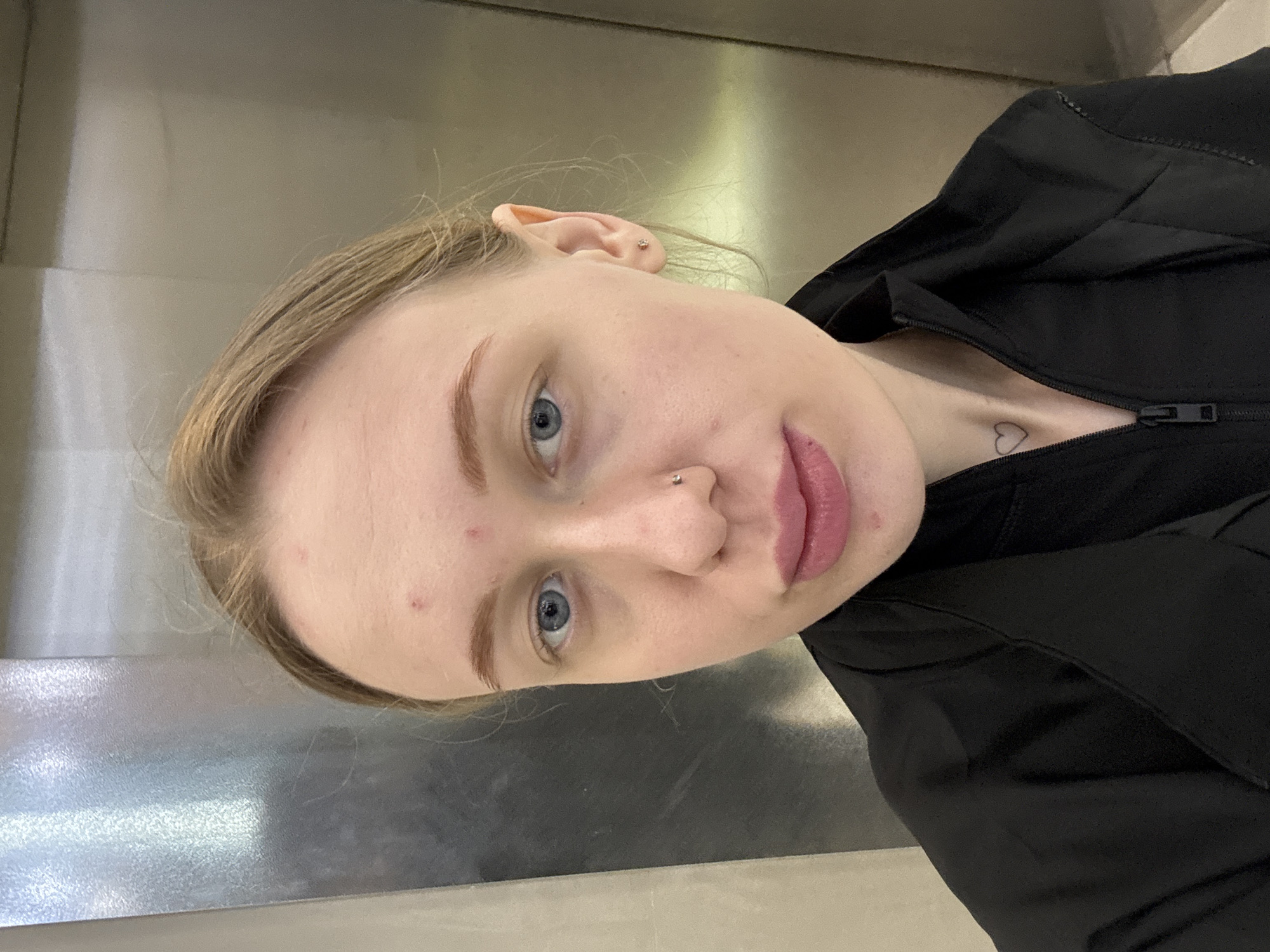 A fair-skinned woman with blonde hair and blue eyes is posing for a selfie. She has a nose piercing, a small heart tattoo on her chest, and is wearing a black jacket. The background appears to be an elevator with a metallic surface. There is no indication of Rostov-on-Don, Russia, in the image.