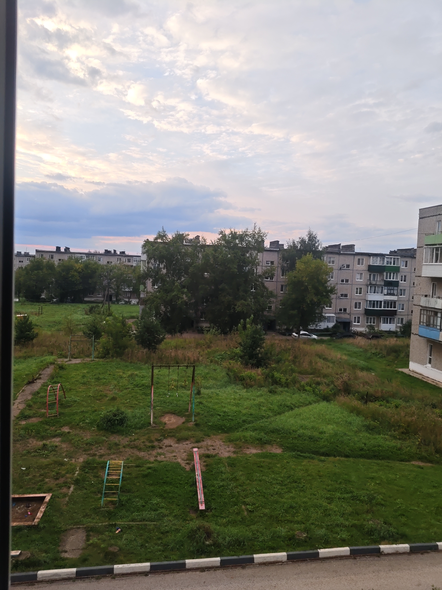The image is an outdoor, elevated view of a residential courtyard in the city of Kurashim, Russia. The scene is dominated by a cloudy sky with soft pink and yellow hues suggesting either sunrise or sunset. In the foreground, a paved area with a black and white curb separates the courtyard from a road. The courtyard itself is a grassy area with patches of dried vegetation and several pieces of playground equipment. These include a sandbox with some debris, a ladder, a swing set with two swings, and a long, pink slide. Several trees and bushes are scattered throughout the courtyard, with a line of tall apartment buildings visible in the background. The buildings are made of concrete and appear to be of Soviet-era construction, with multiple stories and balconies. A white car is parked in front of one of the buildings. The overall impression is one of a somewhat weathered but functional urban environment.