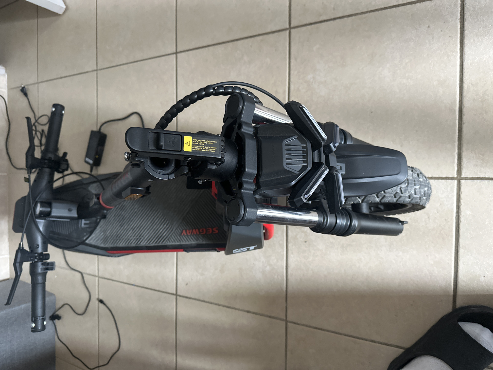 This image shows a close-up, top-down view of a black and red electric scooter, identified by the "SEGWAY" logo visible on its deck. The scooter is parked on a tiled floor, which appears to be indoors, possibly in a garage or hallway, given the presence of a white cabinet or shelf in the upper left corner. The scooter is not in use; its handlebars are folded down, and power cables for charging are connected and visible.
The scooter has a rugged design with thick tires, suggesting it's built for varied terrain. The handlebars are equipped with grips, levers, and what appears to be a display unit. A yellow warning sticker with text in English and French is attached to the handlebar mechanism, reading "Make sure the safety button is in the locked position before riding." Other visible text includes "OPEN".
No people are visible in the image, and the lighting suggests it is daytime. The overall scene is static, focusing on the details of the electric scooter and its charging accessories. The tiled flooring provides a neutral background that emphasizes the scooter itself. There are no specific weather cues, and the location context of Anduze, France, doesn't offer any visual clues within the image itself. The image is a straightforward depiction of an object in its environment, with a focus on technical details and safety information.
