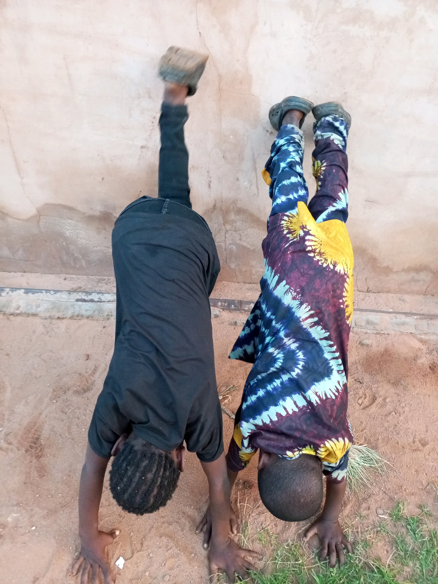 Two young boys are performing handstands against a textured, beige wall. The boy on the left wears a black t-shirt and black pants. He holds a rough, light-colored brick in his right hand, which is extended upwards. The boy on the right is dressed in a vibrantly colored, tie-dye t-shirt featuring patterns of blue, white, purple, and yellow. He wears dark clogs on his feet. Both boys have their legs extended upwards, away from the wall. The ground beneath them is composed of sandy soil with patches of dry grass and some sparse green vegetation on the right side. The overall scene appears to be outdoors, possibly in a yard or open area. The image is taken from a slightly elevated angle looking down.
